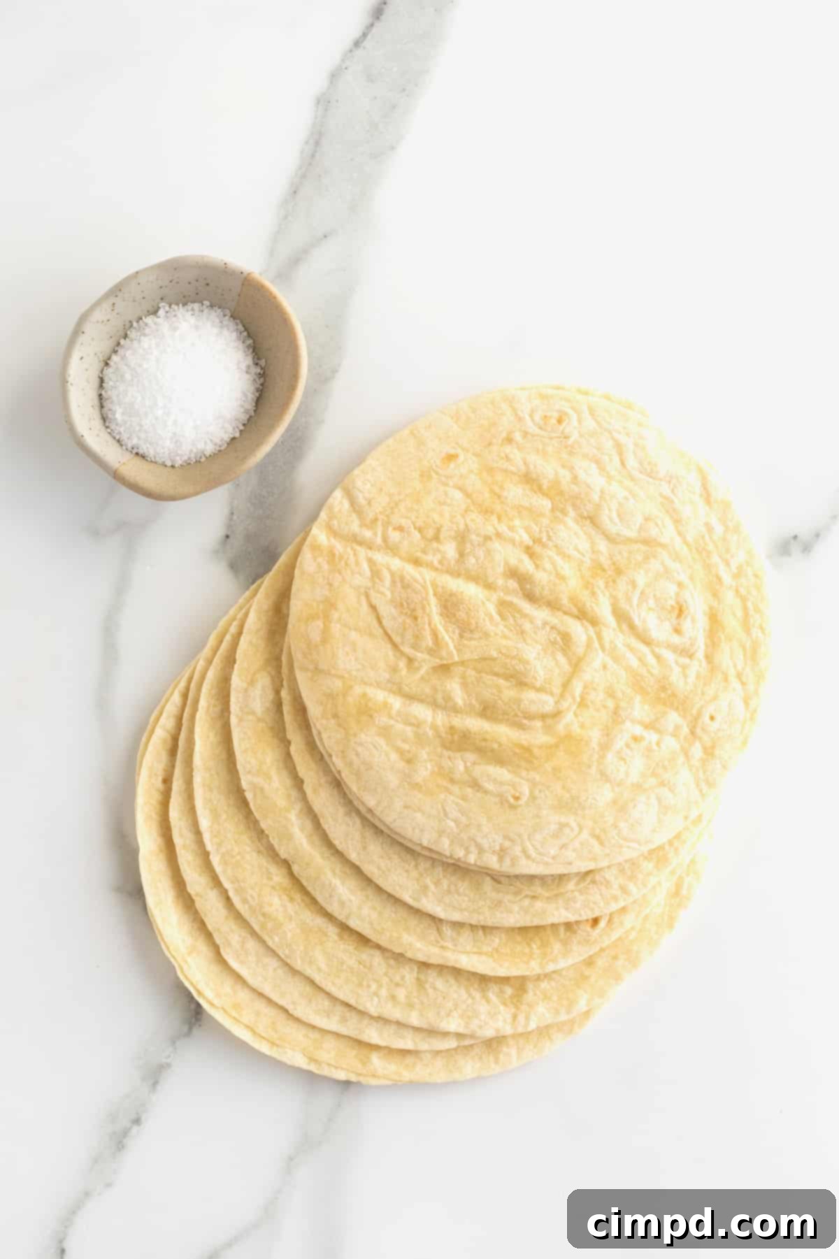 A stack of soft whole flour tortillas placed next to a small white dish filled with sparkling kosher salt.