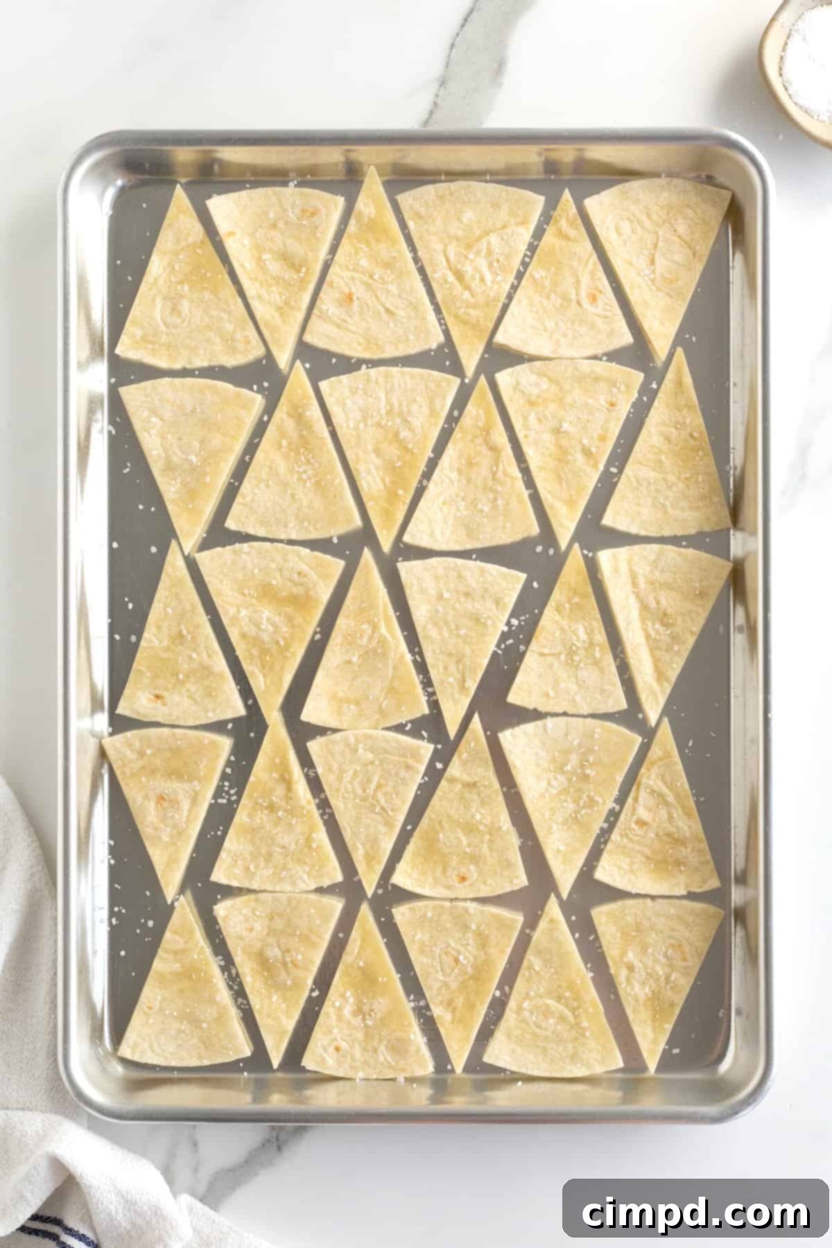 Triangle-cut flour tortillas meticulously arranged on a baking sheet, glistening lightly with cooking spray and evenly sprinkled with coarse sea salt.