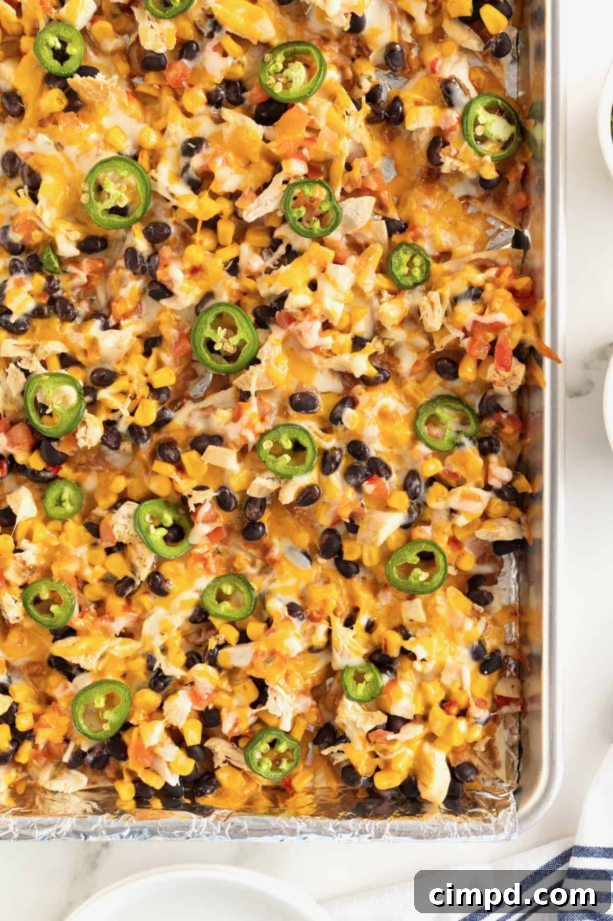 Loaded One Pan Chicken Black Bean Nachos 2 An aluminum sheet pan filled with golden-brown nachos topped generously with melted cheese, vibrant black beans, and bright jalapeño slices.
