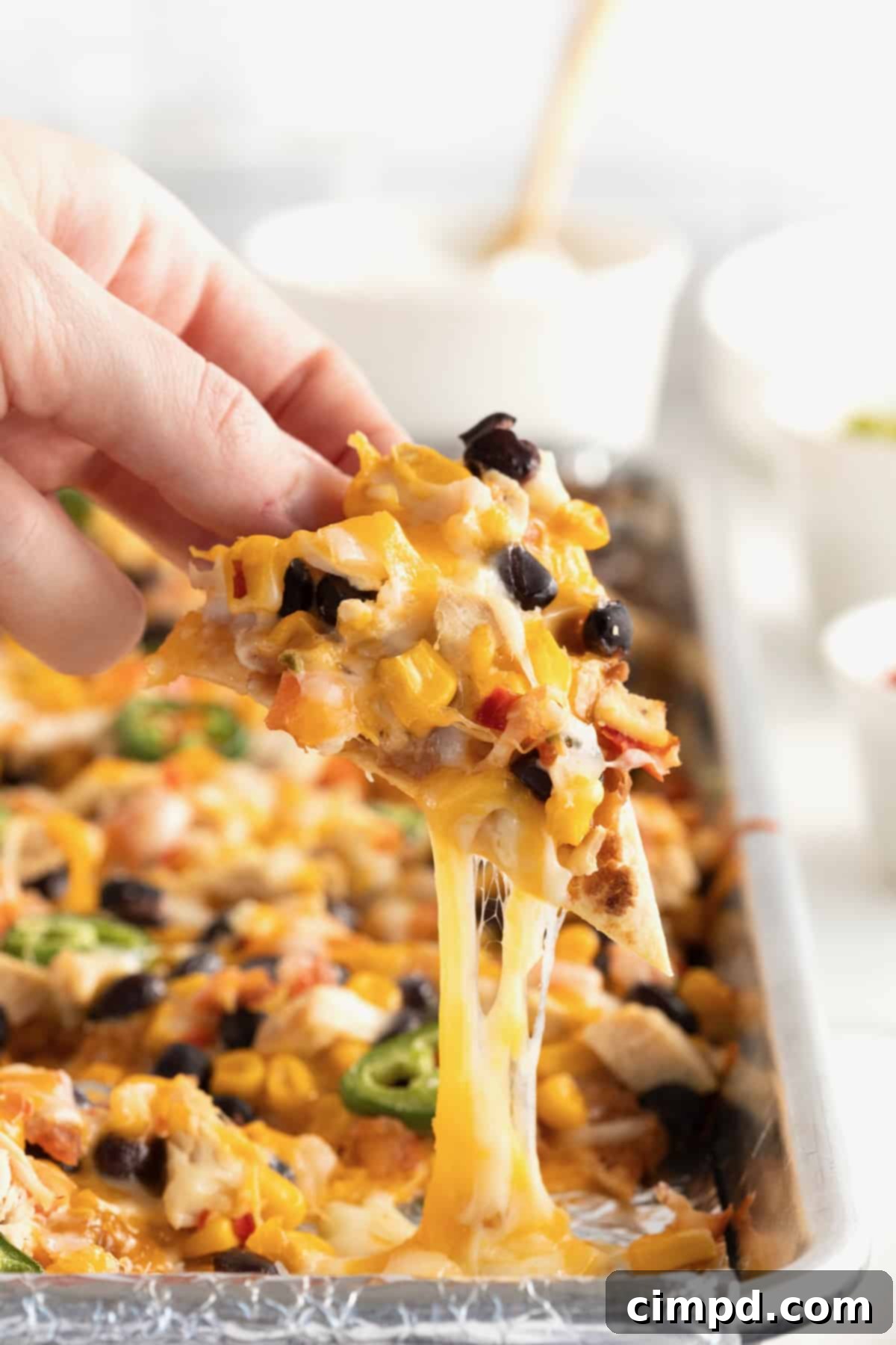 Loaded One Pan Chicken Black Bean Nachos 3 A perfectly loaded chicken and black bean nacho, adorned with melted cheese and fresh toppings, being carefully lifted from an aluminum sheet pan.
