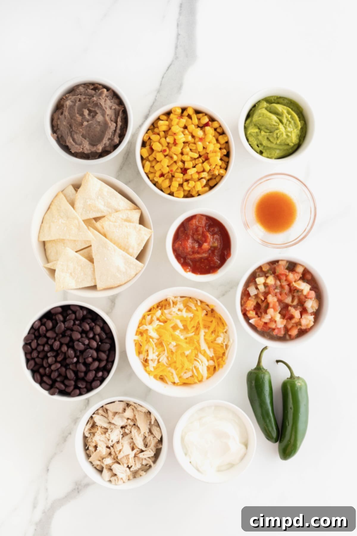 Loaded One Pan Chicken Black Bean Nachos 5 A beautiful arrangement of fresh ingredients for chicken and black bean nachos, presented in small white glass dishes on a pristine white marble counter, with two vibrant jalapeño peppers in the foreground.