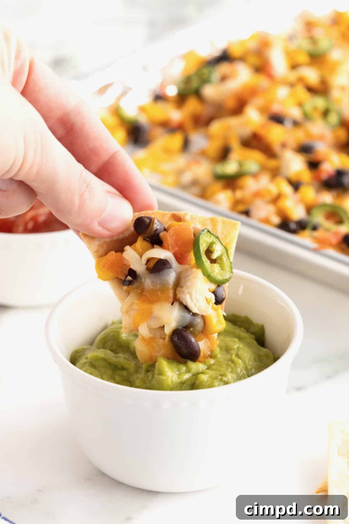 Loaded One Pan Chicken Black Bean Nachos 8 A single, perfectly loaded chicken and black bean nacho being dipped into a small, inviting white bowl of creamy guacamole, ready to be enjoyed.