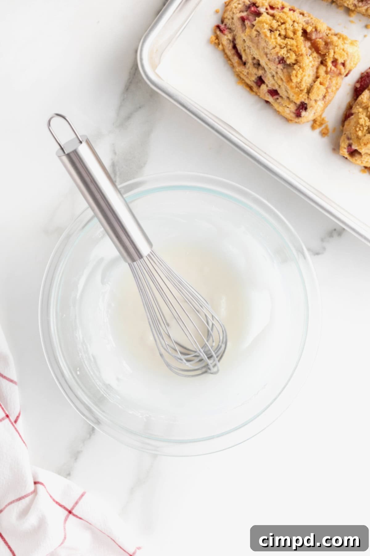 A medium clear mixing bowl containing smooth white icing, with a wire whisk resting inside, ready for drizzling.