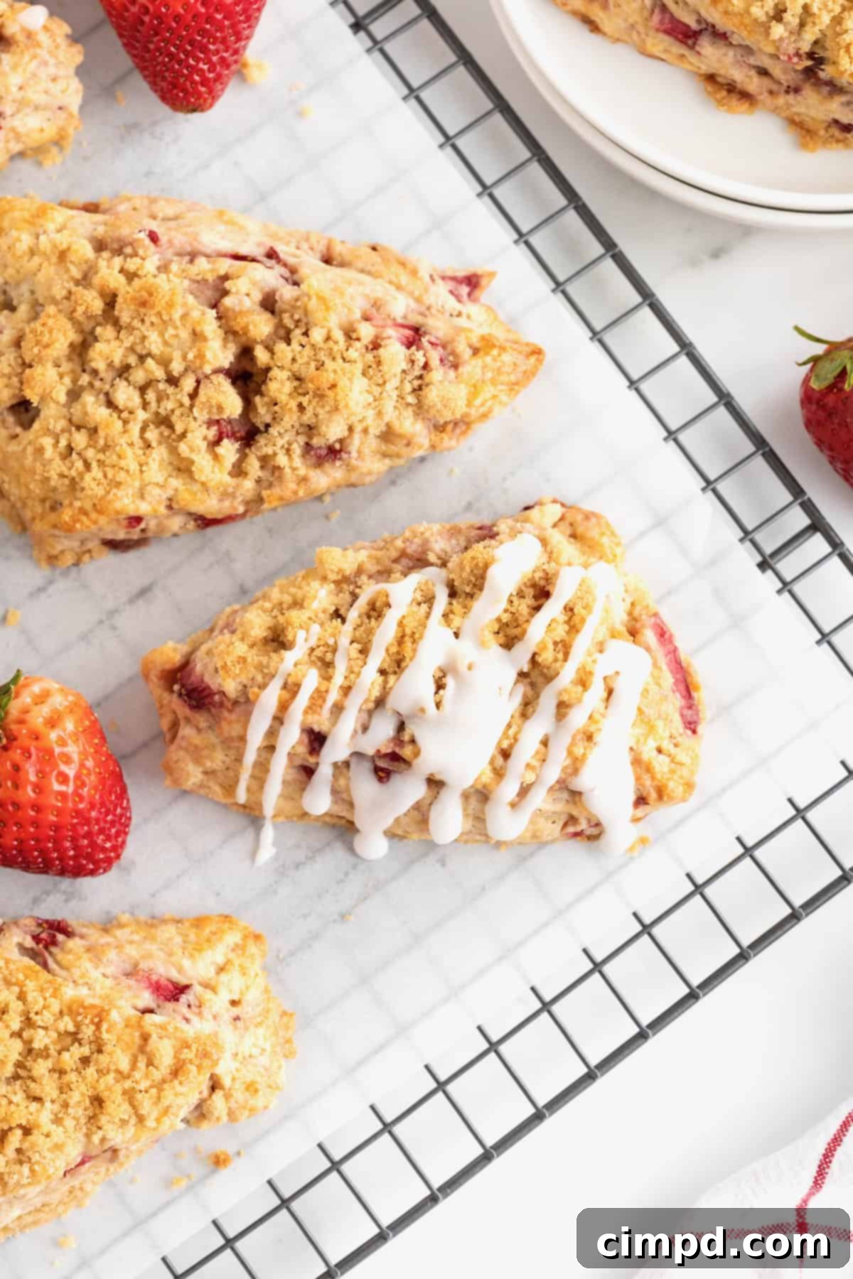 A close-up of a strawberry scone being drizzled with a simple white icing, with two other scones in the background on a cooling rack.