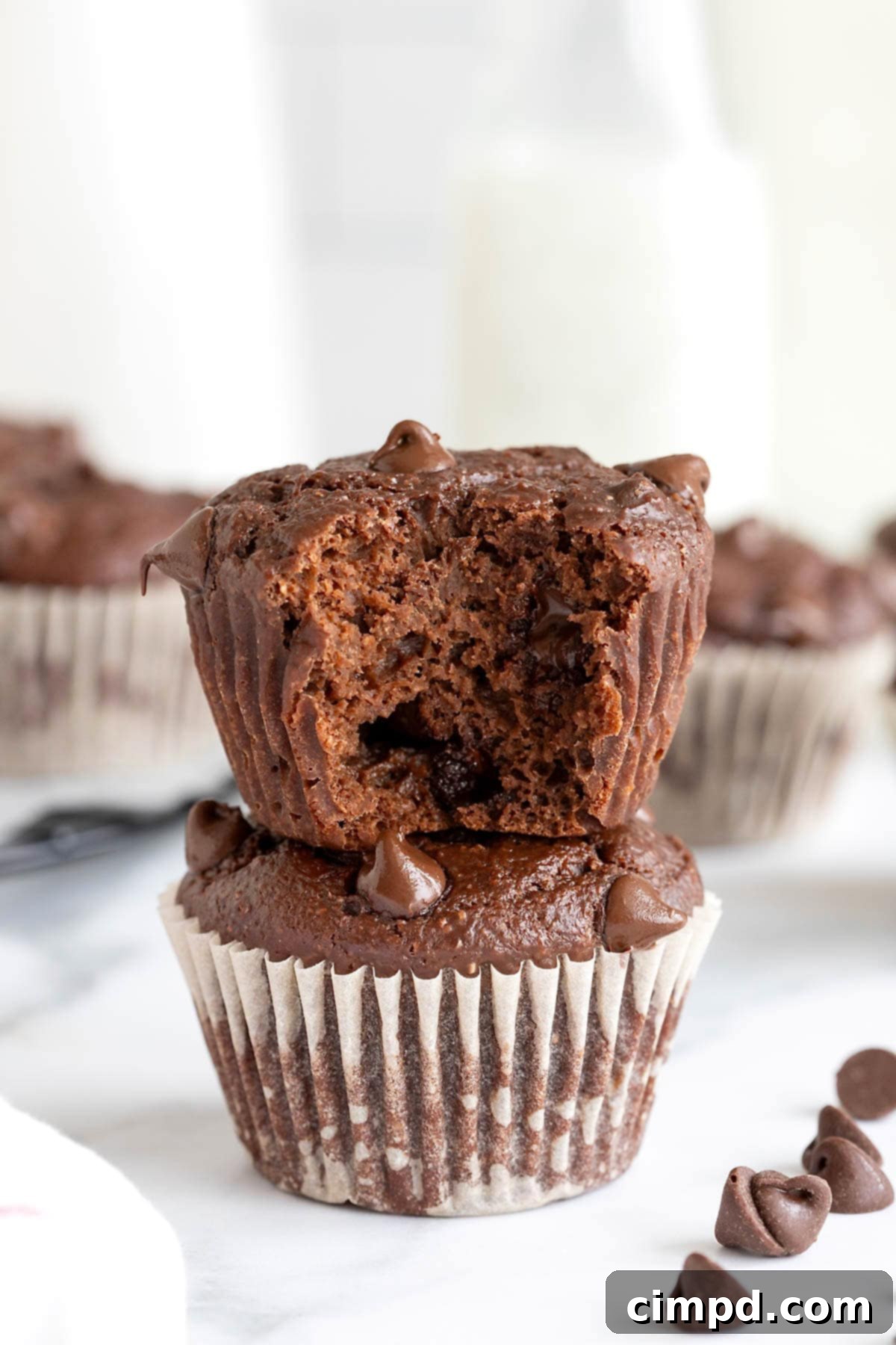 Two irresistibly moist Healthier Double Chocolate Muffins with white paper wrappers, stacked on a clean white marble counter. The top muffin shows a delicious bite taken out, revealing its rich, fluffy interior.