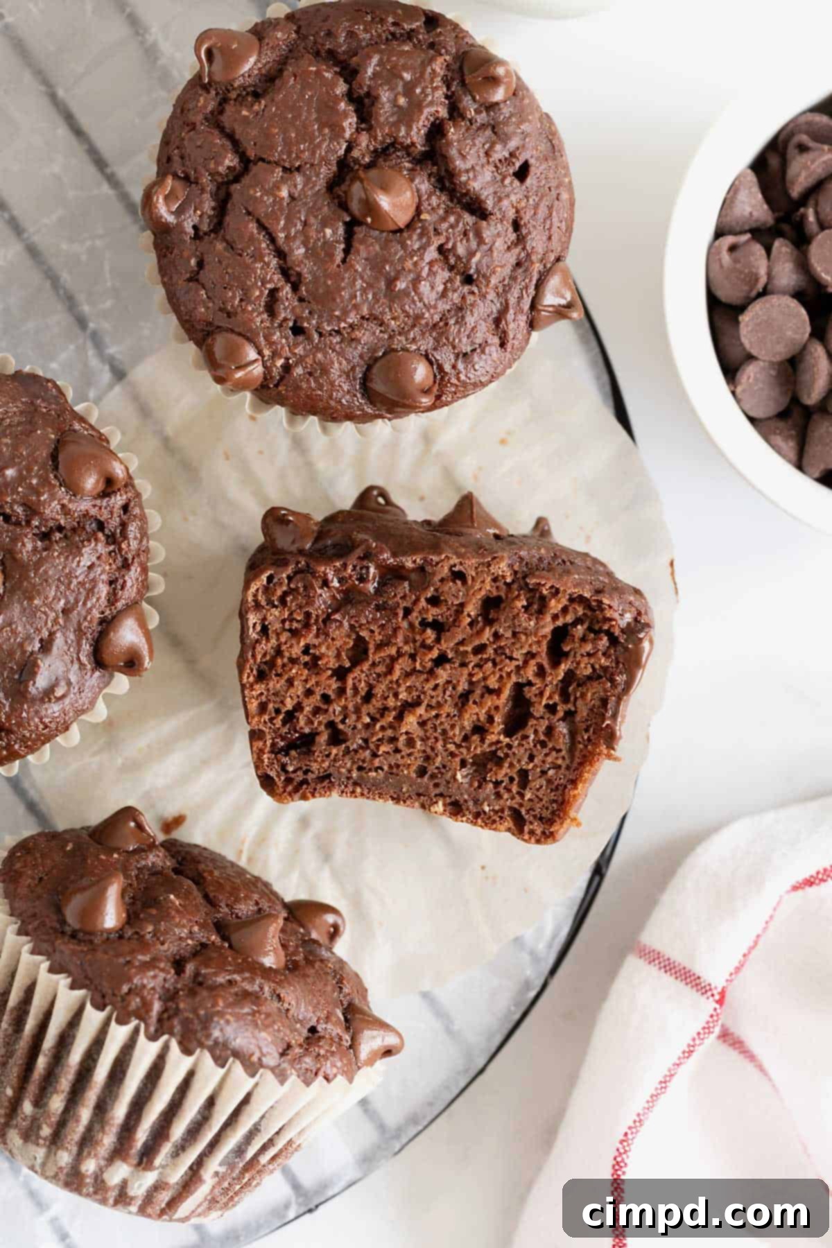 A round wire cooling rack showcases several Healthier Double Chocolate Muffins, with one muffin thoughtfully cut in half to reveal its tender, chocolatey crumb and generous chocolate chip distribution.