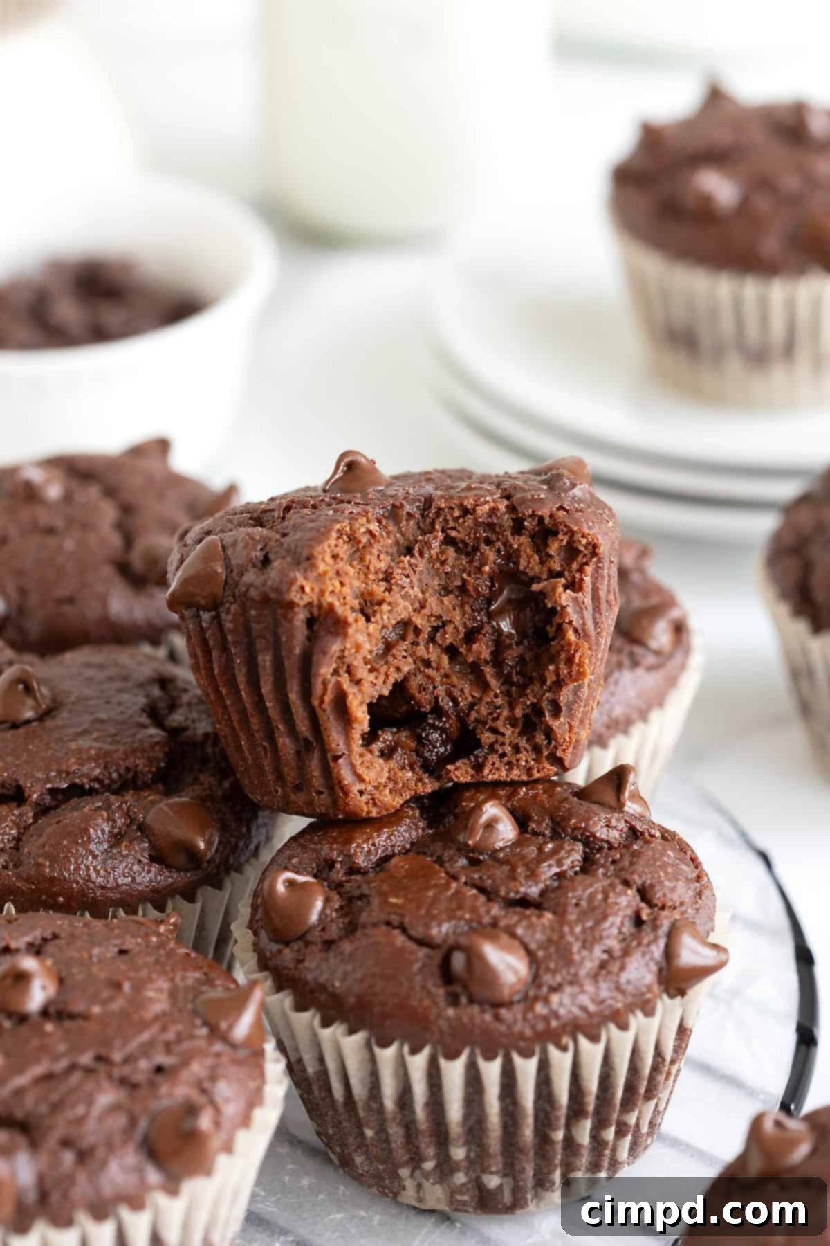 Chocolate muffins piled artfully on a round wire cooling rack. The top muffin enticingly shows a bite taken out, revealing the rich, moist interior and melted chocolate chips.