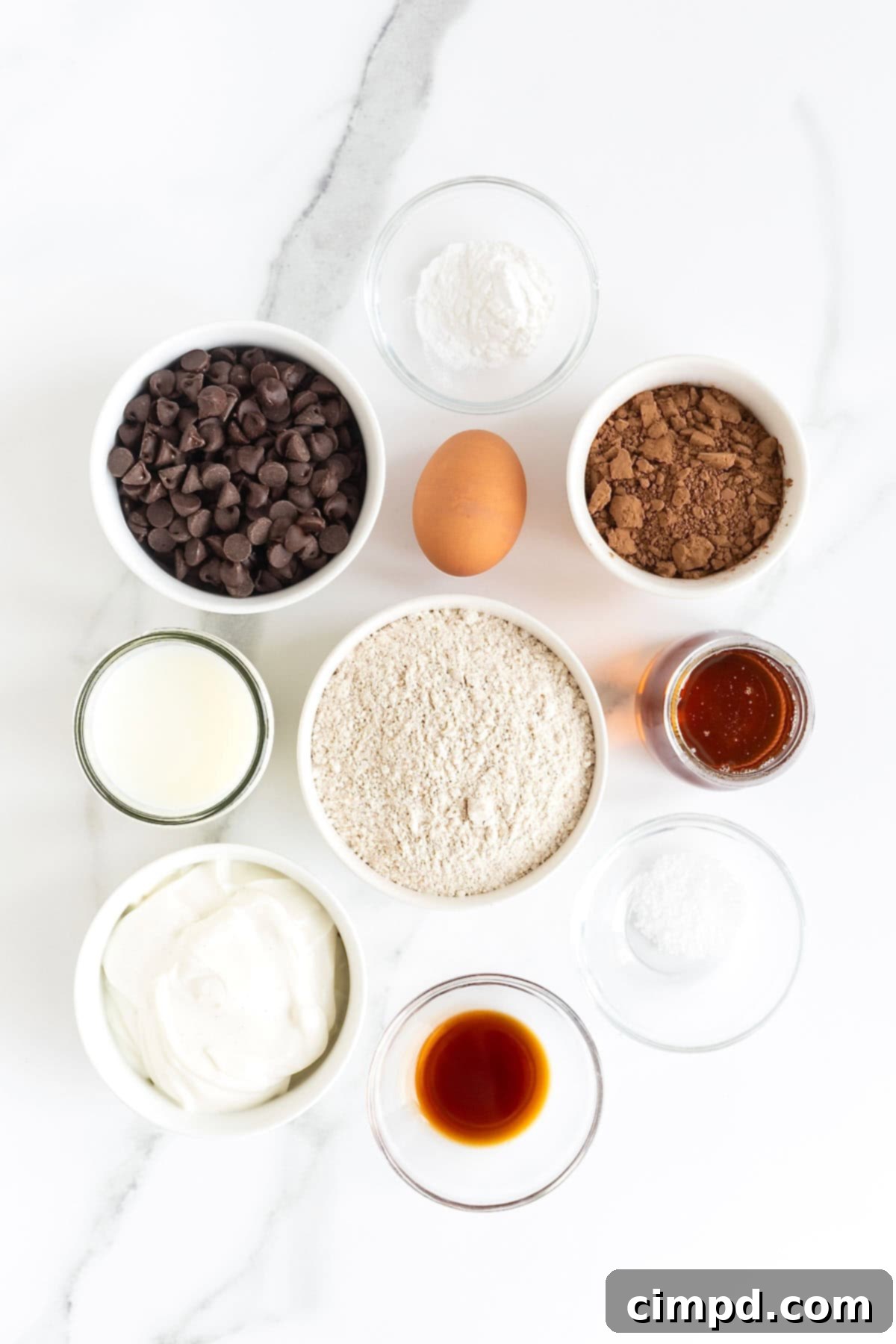 Ingredients for healthier double chocolate muffins meticulously arranged in small white dishes on a pristine white marble counter, ready for baking.