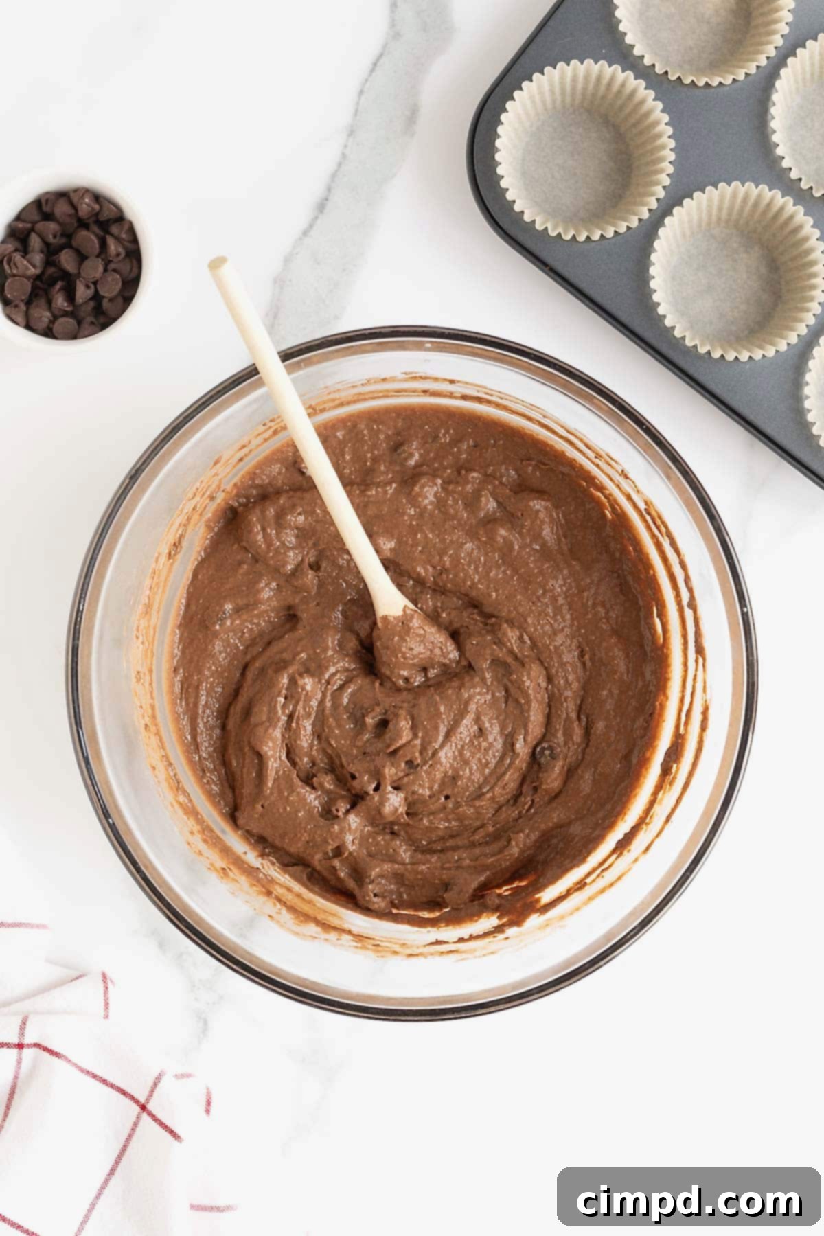 Rich chocolate muffin batter resting in a glass mixing bowl, with a wooden spoon sticking out, signaling it's ready for the next step.