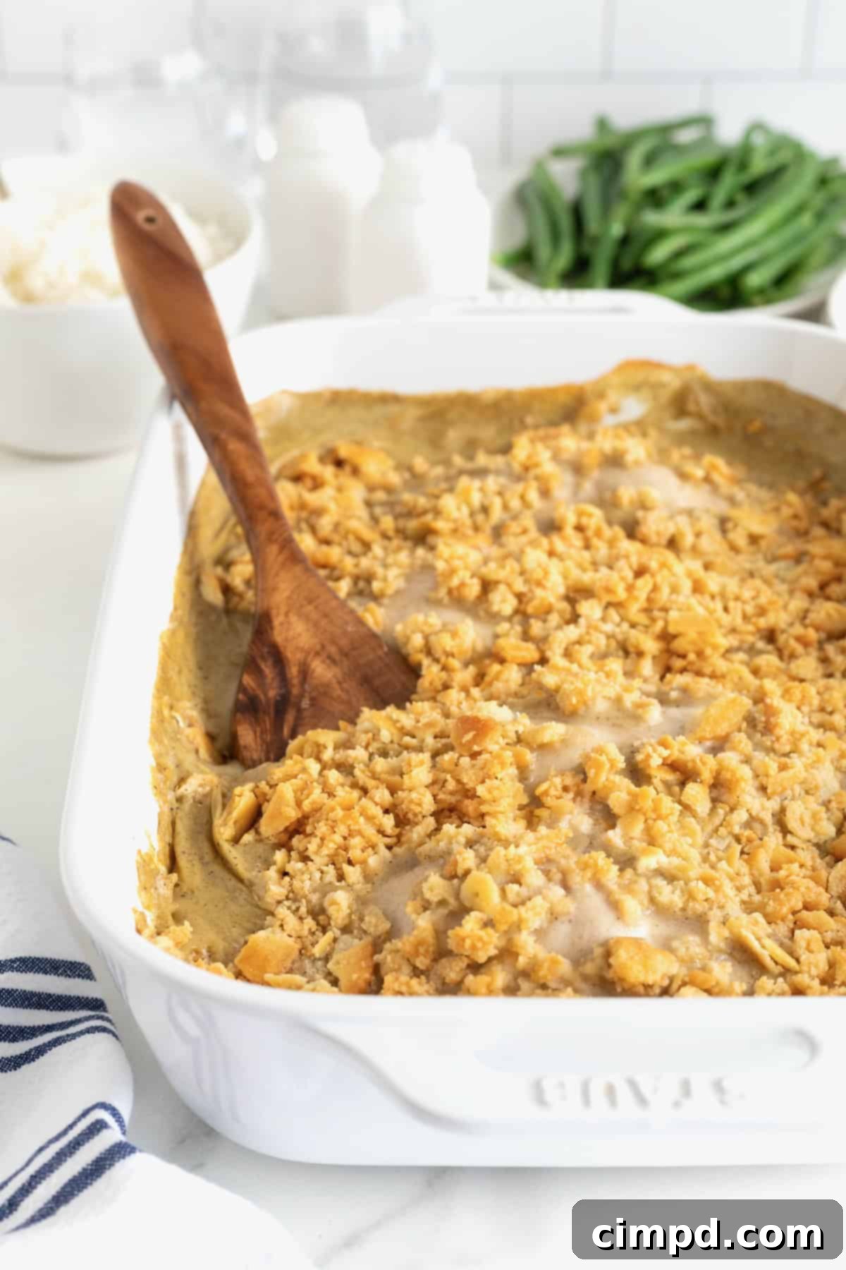 A pristine white baking dish filled with a creamy chicken casserole, topped with golden-brown cracker crumbs, with a wooden spoon ready for serving.