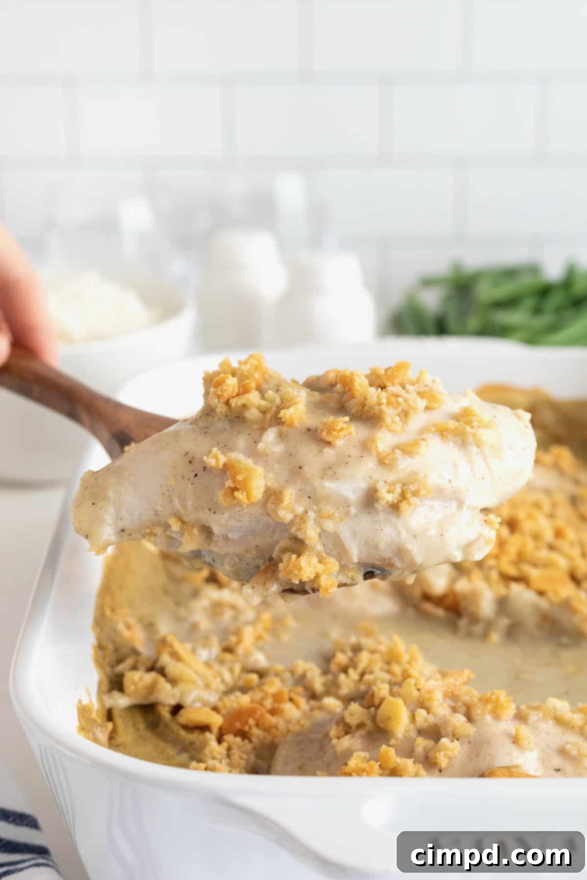 A wooden spoon scooping a portion of creamy chicken bake from a white casserole dish, highlighting the rich sauce and golden cracker topping.