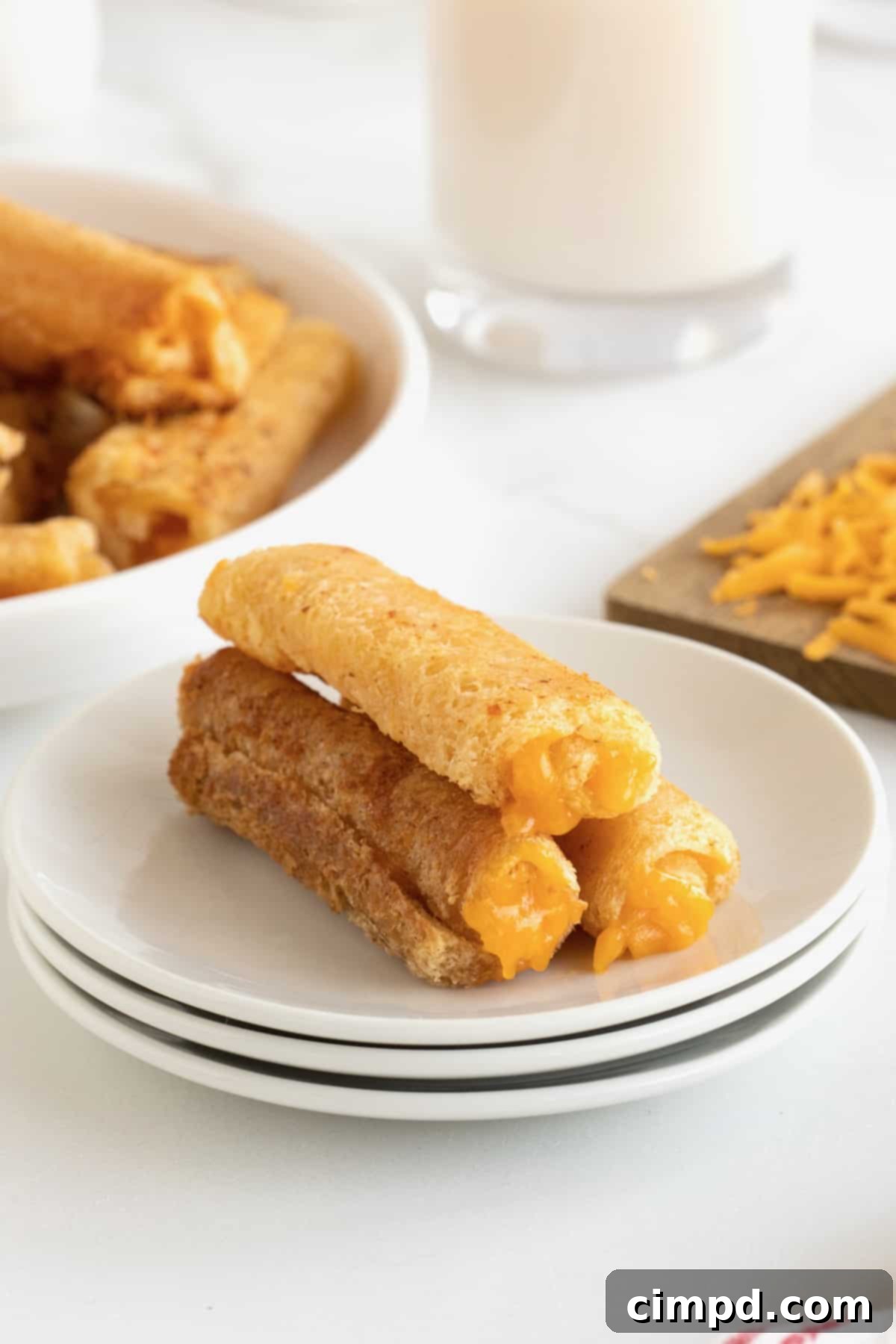 Three perfectly golden-brown grilled cheese roll ups neatly arranged on a white plate, with a wooden cutting board and shredded yellow cheese subtly blurred in the background, hinting at fresh preparation.
