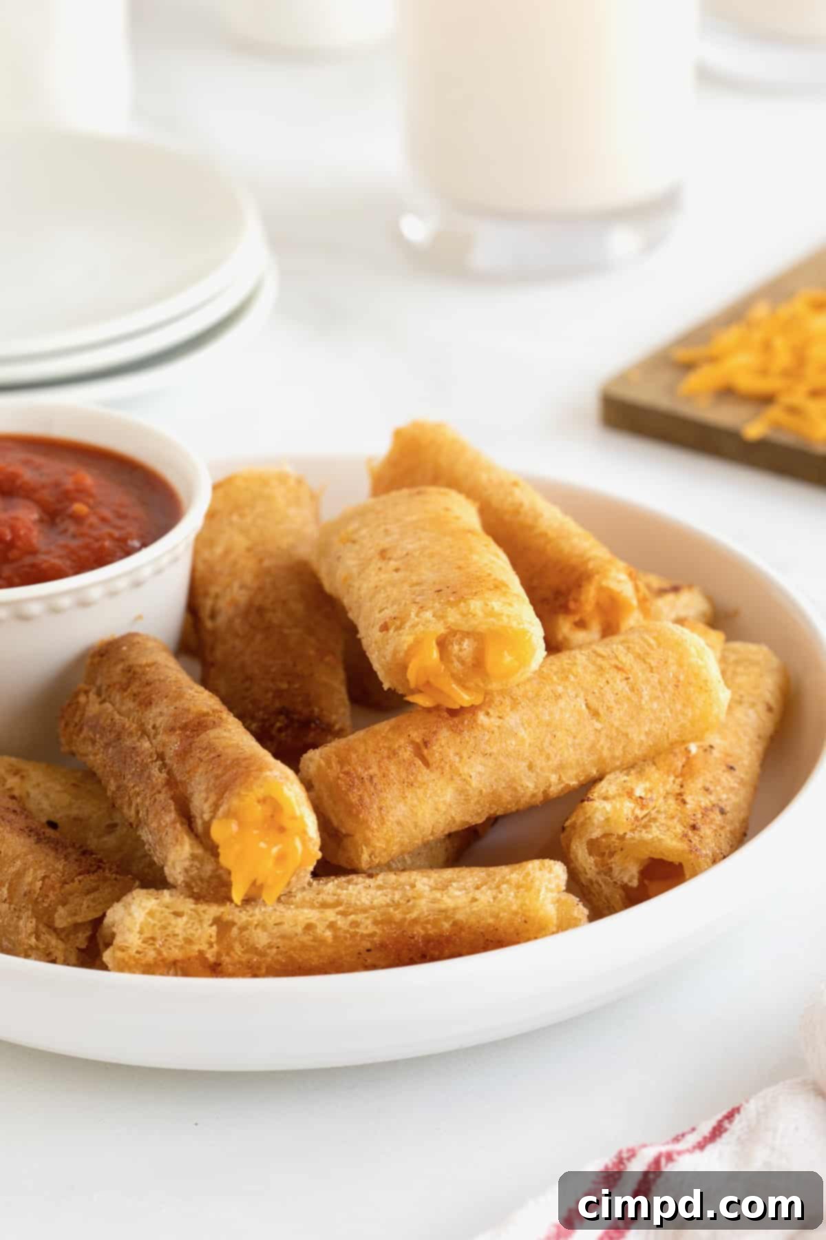 A large white serving plate with an abundant pile of grilled cheese roll ups, accompanied by a small, inviting dish of marinara sauce for dipping.