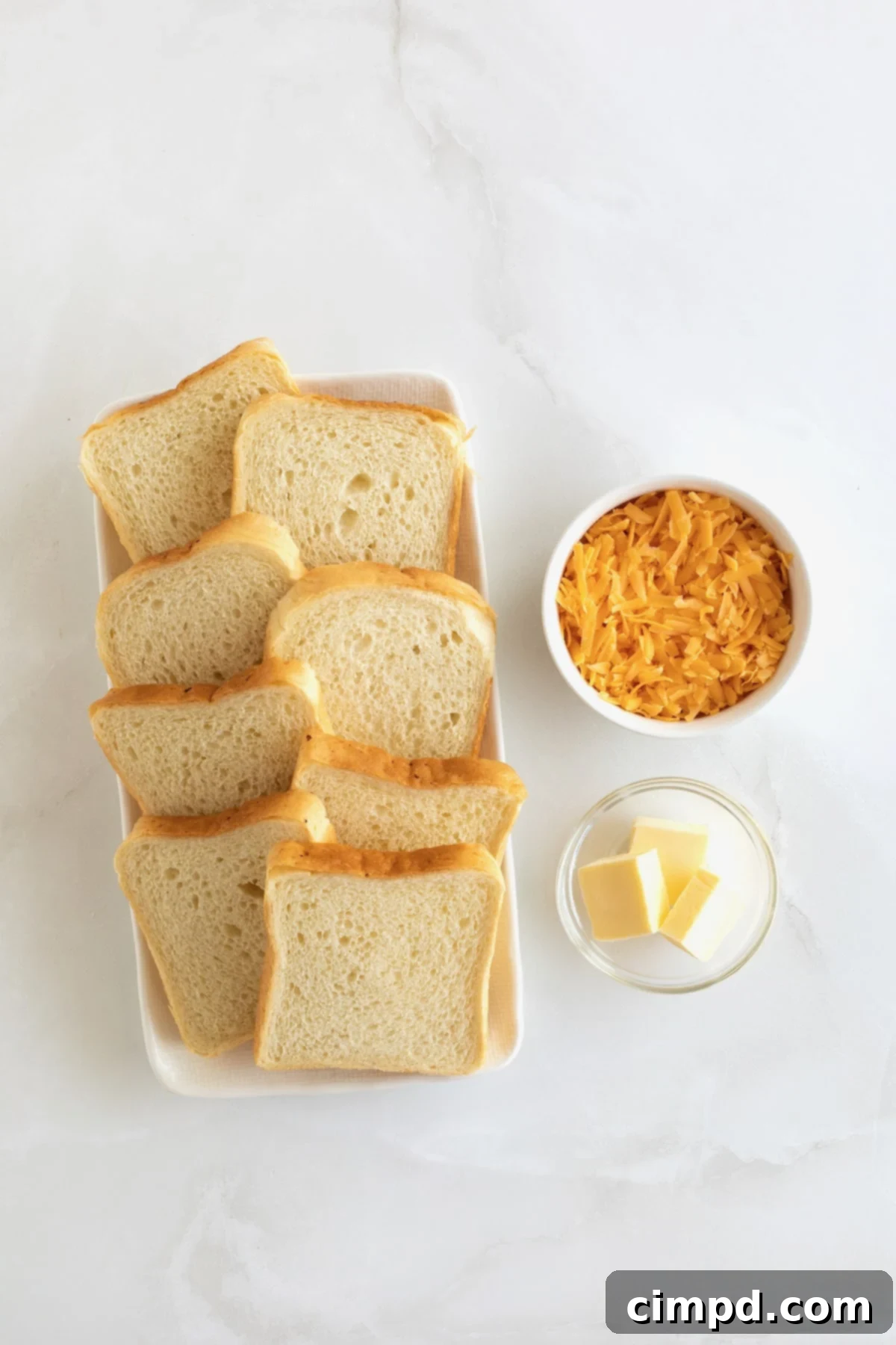 A pristine white marble countertop showcasing all the fresh ingredients meticulously arranged for making grilled cheese roll ups, including bread, shredded cheese, and butter.
