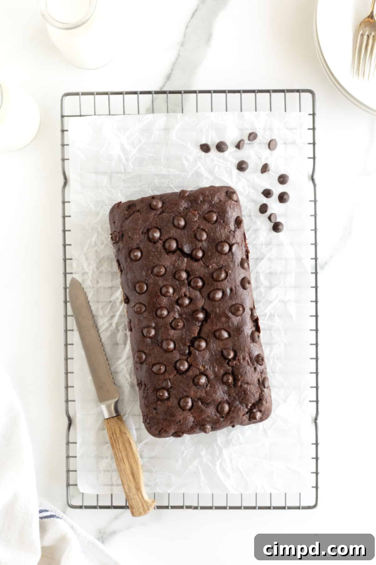 Guilt-Free Chocolate Banana Loaf 4 A freshly baked loaf of chocolate banana bread cooling on a wire rack, sprinkled with chocolate chips and accompanied by a bread knife.