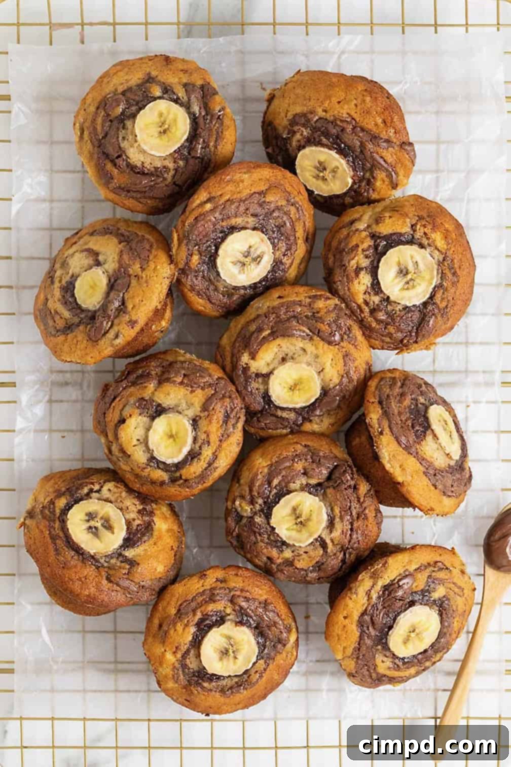 Nutella swirled banana muffins with a slice of banana on top resting on a parchment lined gold tone cooling rack.