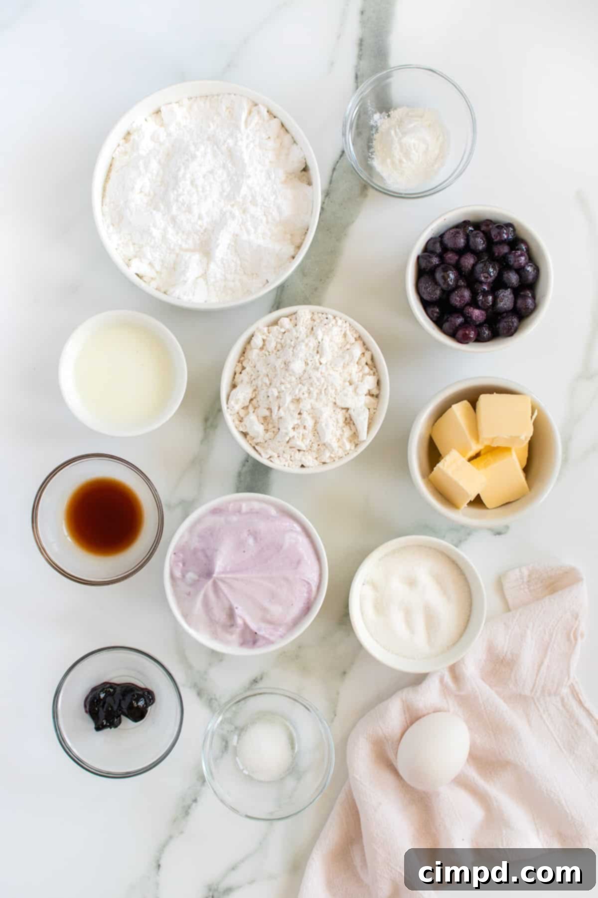 Oven-Baked Blueberry Fritter Poppers 4 A collection of ingredients for blueberry fritters, neatly arranged in small glass bowls on a white marble surface.