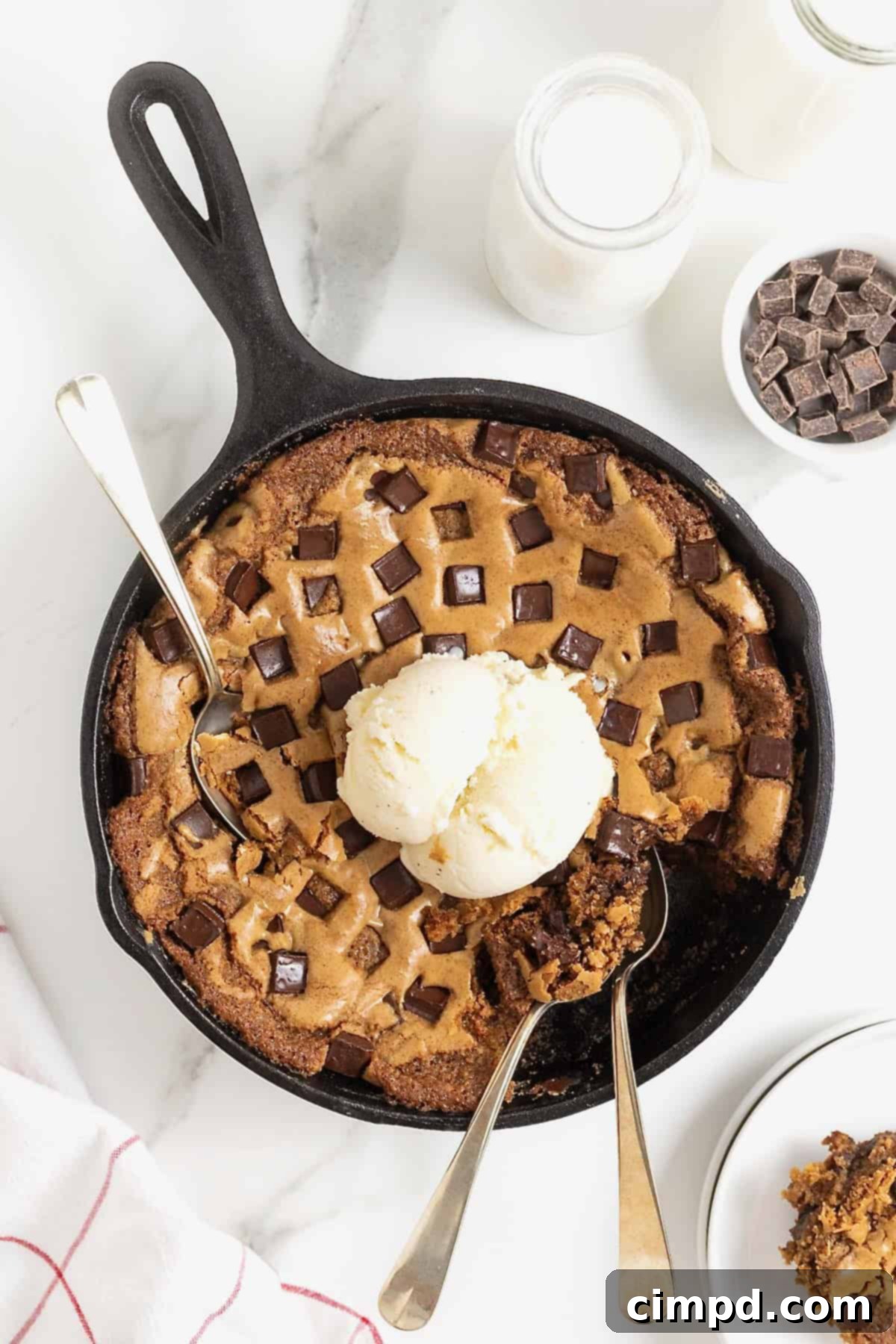 A chocolate chunk cookie in a large black skillet. There's a scoop of ice cream on top and three spoons stuck in it.