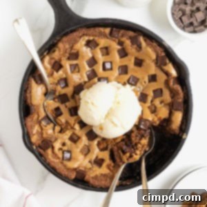 A chocolate chunk cookie in a large black skillet. There's a scoop of ice cream on top and three spoons stuck in it.