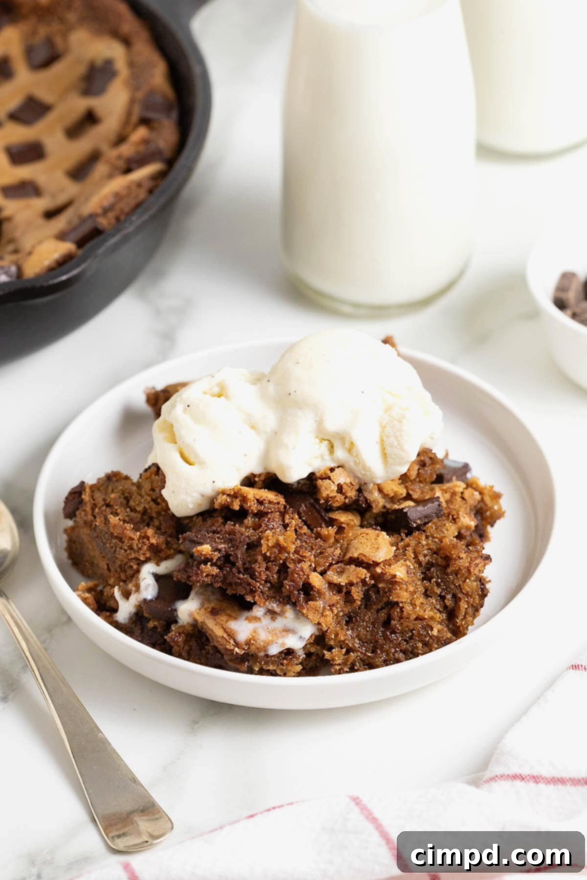 A serving of skillet cookie on a white rimmed plate topped with a scoop of vanilla ice cream.