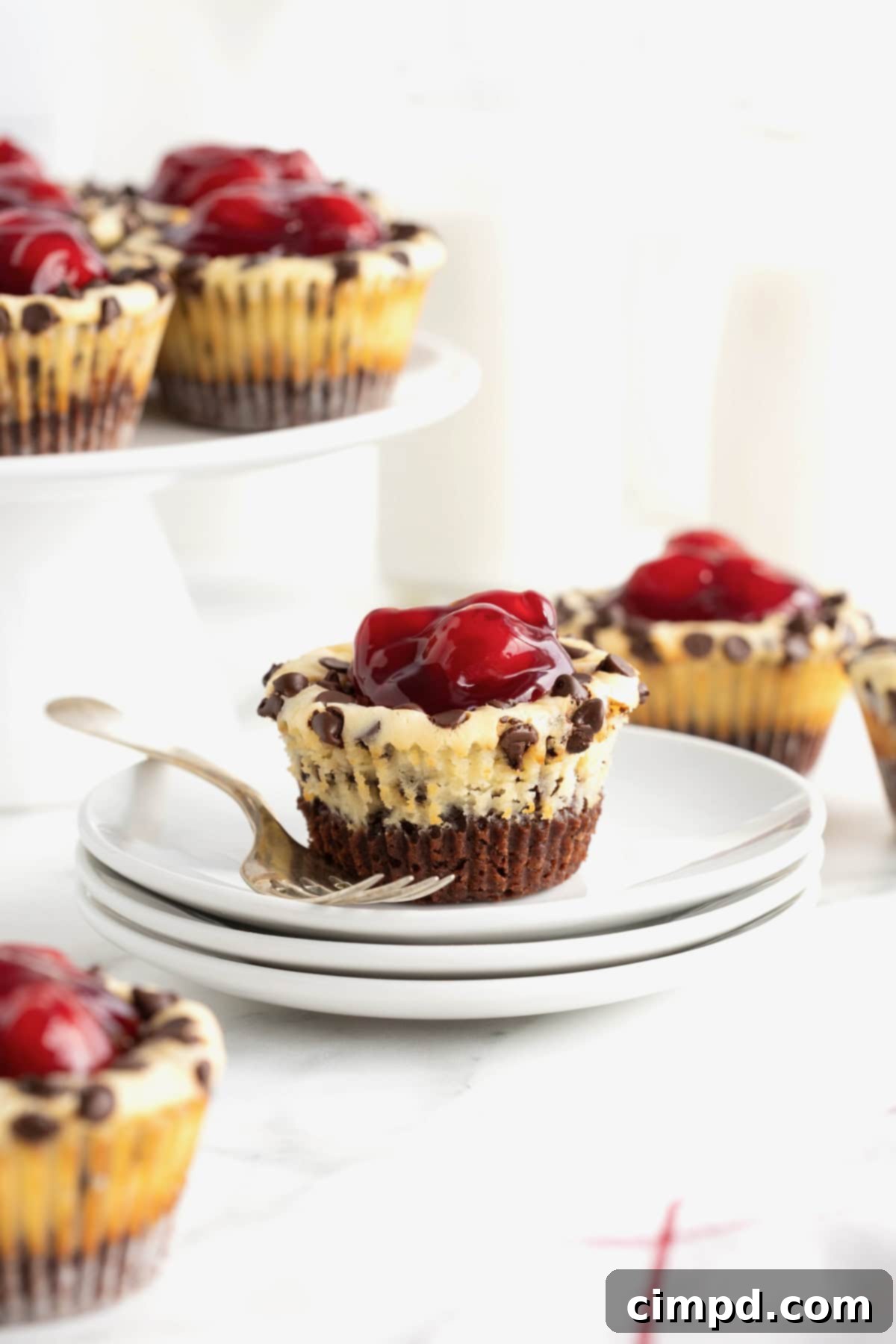 A perfectly portioned Cherry Chocolate Chip Cheesecake Brownie Bite, garnished with a bright red cherry, resting on a stack of pristine white dessert plates.