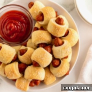 Sausage Swaddles 11 A large white serving plate filled with golden-brown pigs in a blanket, encircling a small glass dish of vibrant red ketchup. The perfect snack for any gathering.