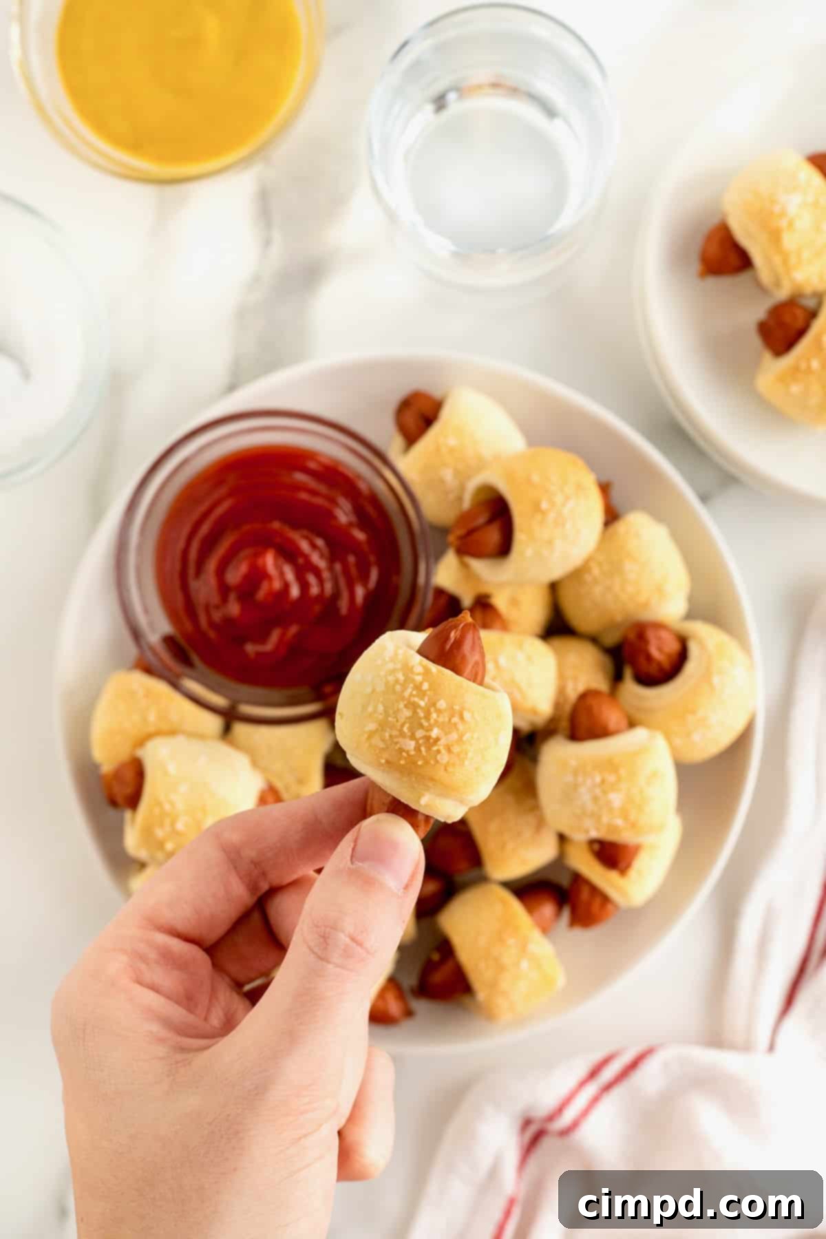 Sausage Swaddles 3 A hand reaches into a large white serving plate filled with delicious pigs in a blanket and a small dish of ketchup, picking up a single golden-brown snack.