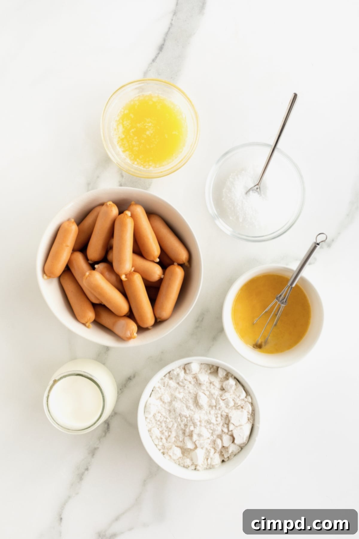Sausage Swaddles 5 A pristine white marble counter showcases small white dishes holding ingredients for pigs in a blanket, including flour, milk, butter, and mini sausages.
