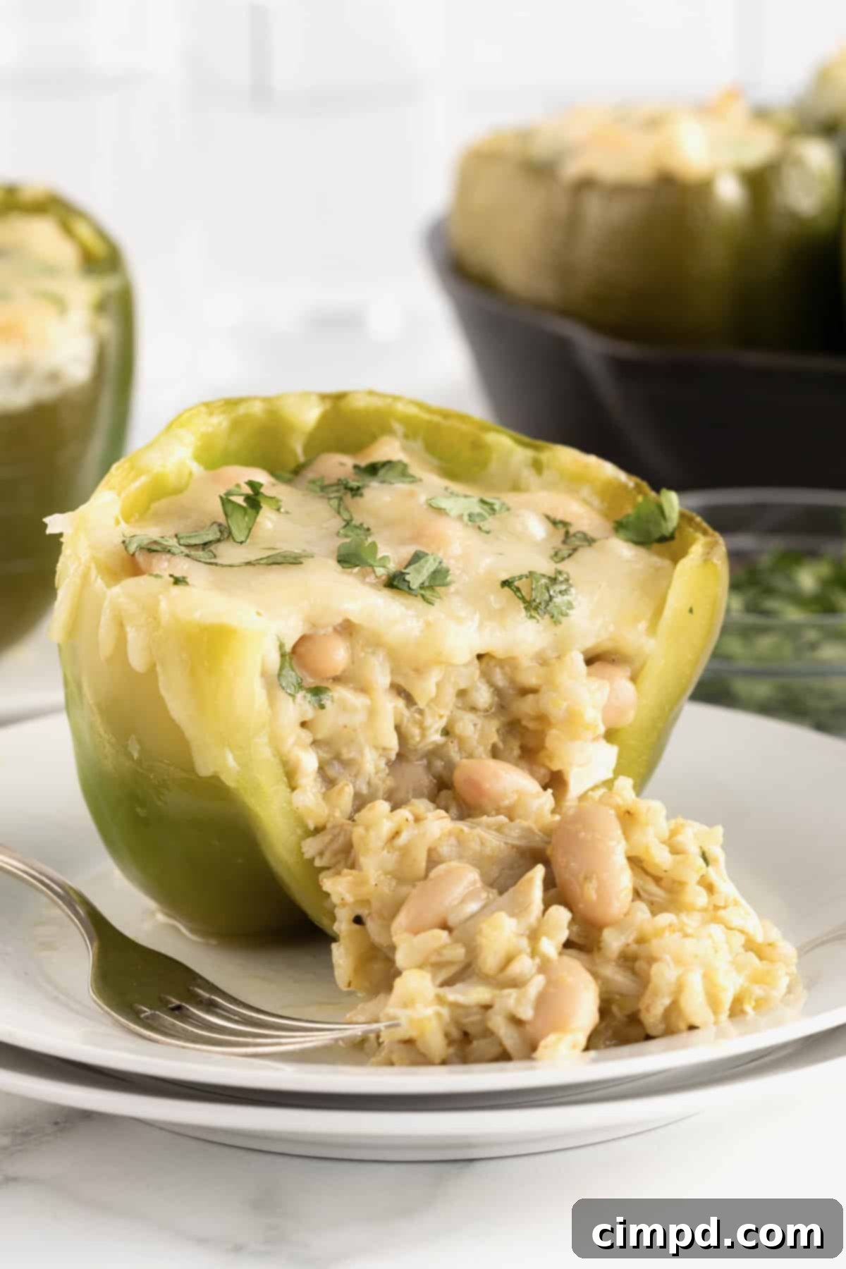 A vibrant green bell pepper, generously stuffed with a creamy chicken, rice, and white bean filling, topped with golden-brown melted white cheese. The pepper is served on a small white plate with a fork resting beside it, highlighting its readiness to be enjoyed.