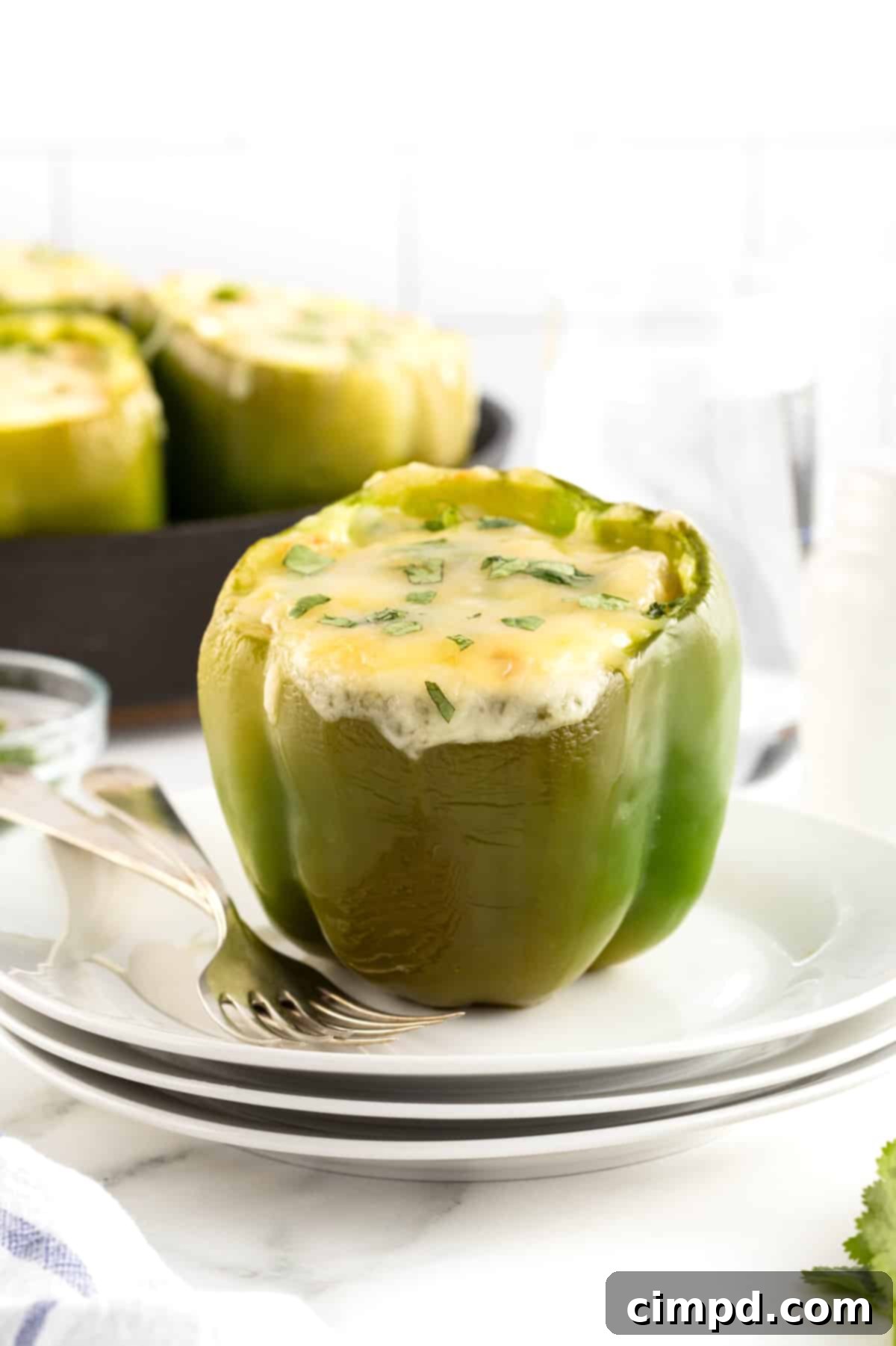 A close-up shot of a single green bell pepper, beautifully stuffed with white chicken chili and topped with melted white cheese and a sprinkle of fresh cilantro, presented on a white plate. A fork is placed elegantly to the left, with a cast iron skillet containing more stuffed peppers blurred in the background, creating a cozy and inviting meal scene.
