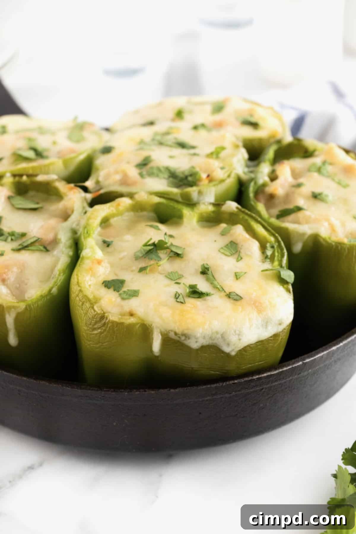 Six vibrant green bell peppers, beautifully stuffed with a hearty white chicken chili mixture and generously topped with melted white cheese. A sprinkle of fresh chopped cilantro adds a touch of color and freshness. The peppers are cozily arranged in a cast iron skillet, ready to be served, set against a clean white background.