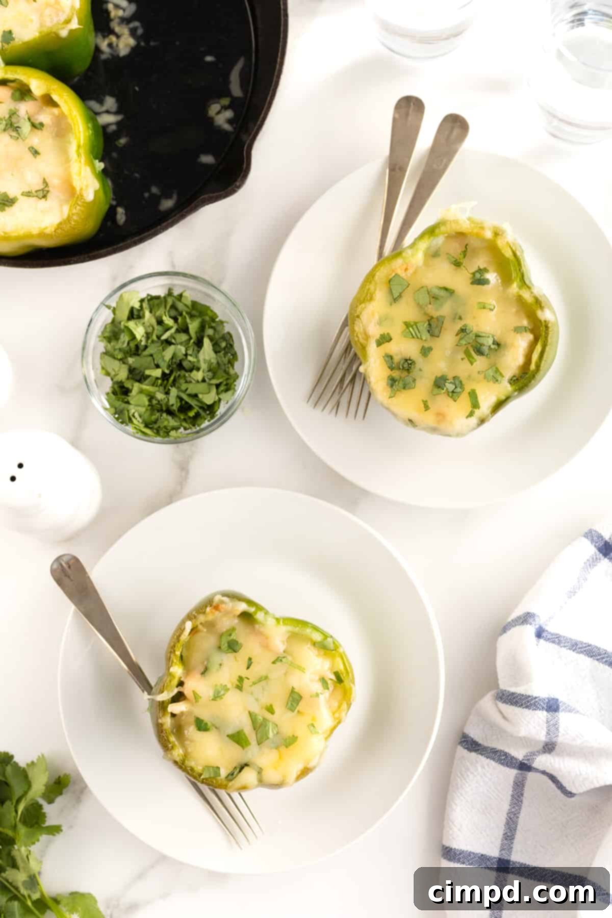Two perfectly cooked green bell peppers, richly stuffed and topped with melted white cheese, are elegantly presented on individual white plates. In the upper left background, a cast iron skillet contains several more stuffed peppers, suggesting a generous meal ready to be shared or enjoyed by a larger family.