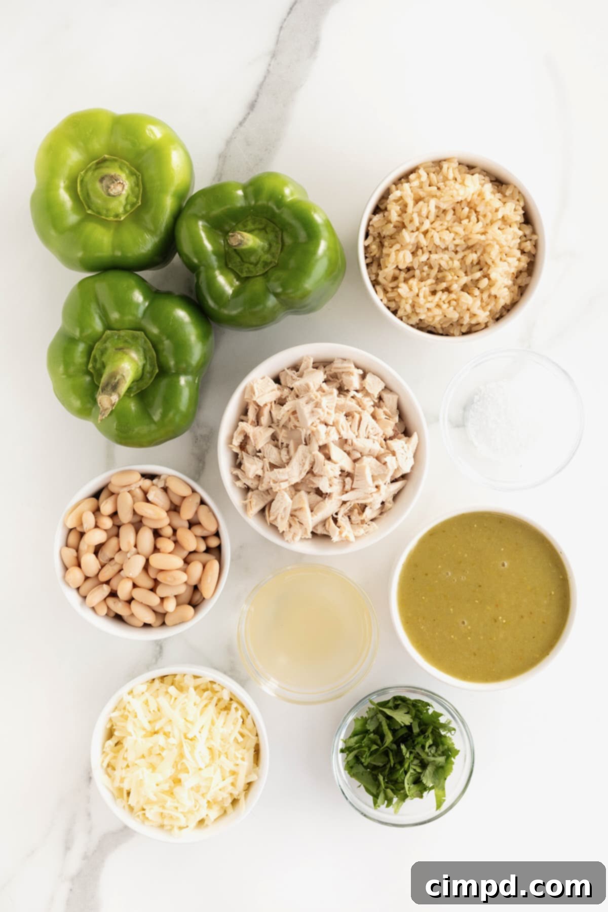A beautifully arranged spread of fresh ingredients for White Chicken Chili Stuffed Peppers on a white marble counter. Three vibrant green bell peppers are prominent, surrounded by small white dishes containing shredded chicken, white beans, cooked quinoa and brown rice, green enchilada sauce, shredded Monterey Jack cheese, and fresh cilantro, all awaiting preparation.