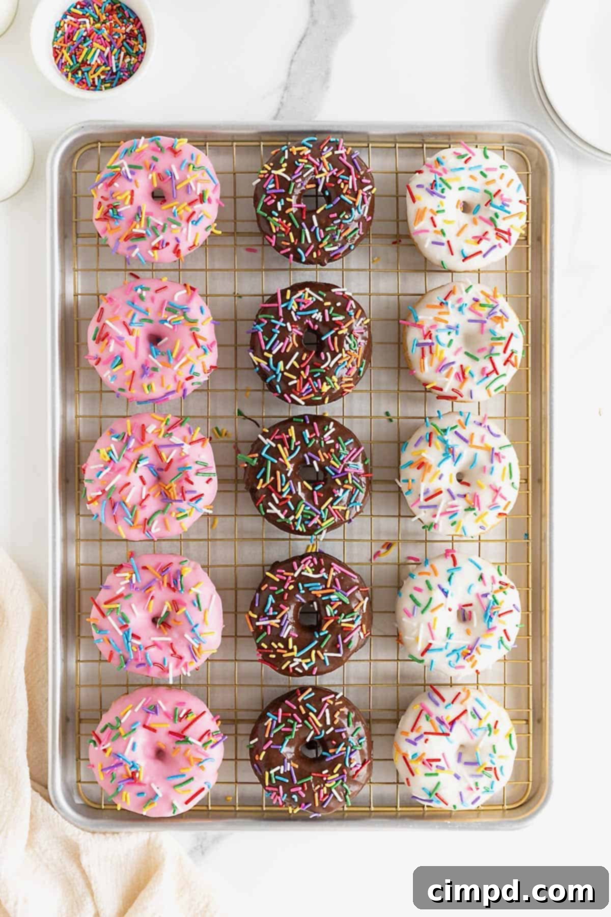 Effortless Cake Mix Baked Donuts 12 A variety of freshly iced strawberry, chocolate, and vanilla baked donuts, each decorated with sprinkles, cooling on a wire rack set within a rimmed baking sheet, collecting any glaze drips.
