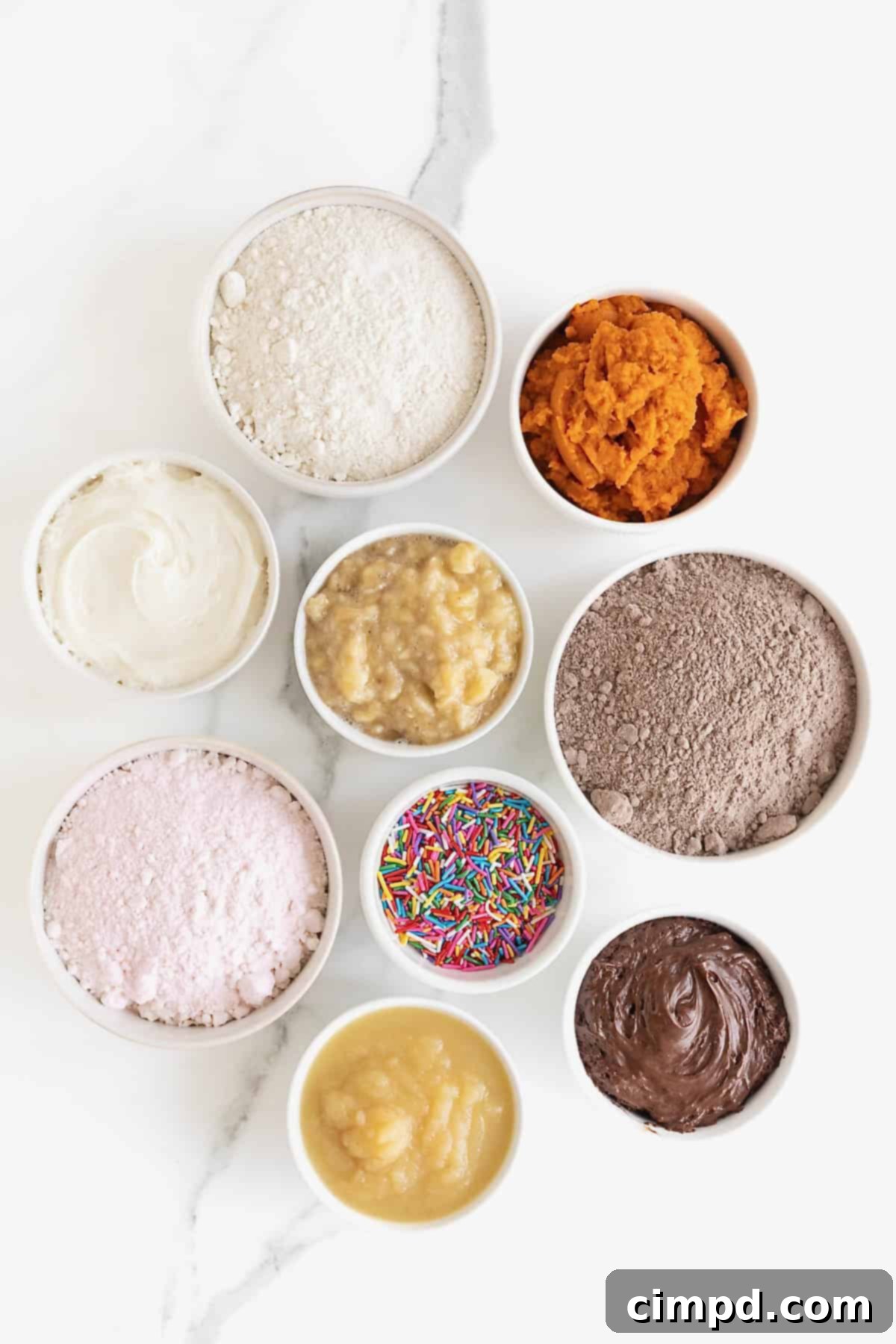 Effortless Cake Mix Baked Donuts 6 Individual ingredients for making cake mix donuts, including a box of cake mix, mashed bananas, and a tub of frosting, neatly displayed in small white dishes on a elegant white marble counter, ready for preparation.