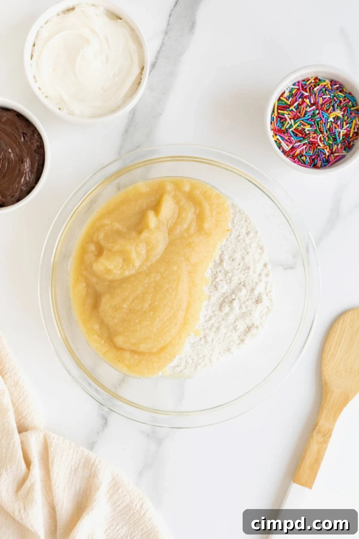 Effortless Cake Mix Baked Donuts 9 Applesauce and dry vanilla cake mix are being combined in a large, clear glass mixing bowl, initiating the simple process of making vanilla cake donuts.