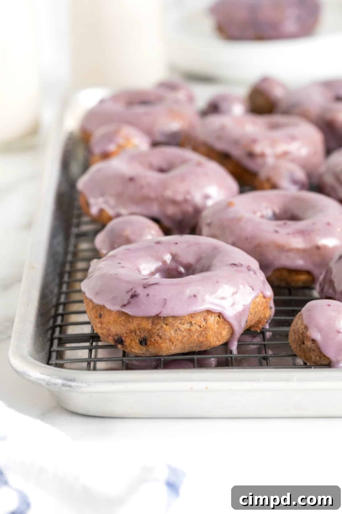 Freshly fried blueberry donuts with a vibrant blueberry glaze cooling on a wire rack, placed over a rimmed baking sheet to catch any drips.