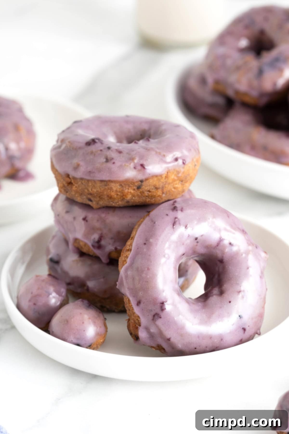 A selection of four blueberry donuts and two small donut holes, all generously coated in a shimmering blueberry glaze, arranged on a pristine white rimmed plate.