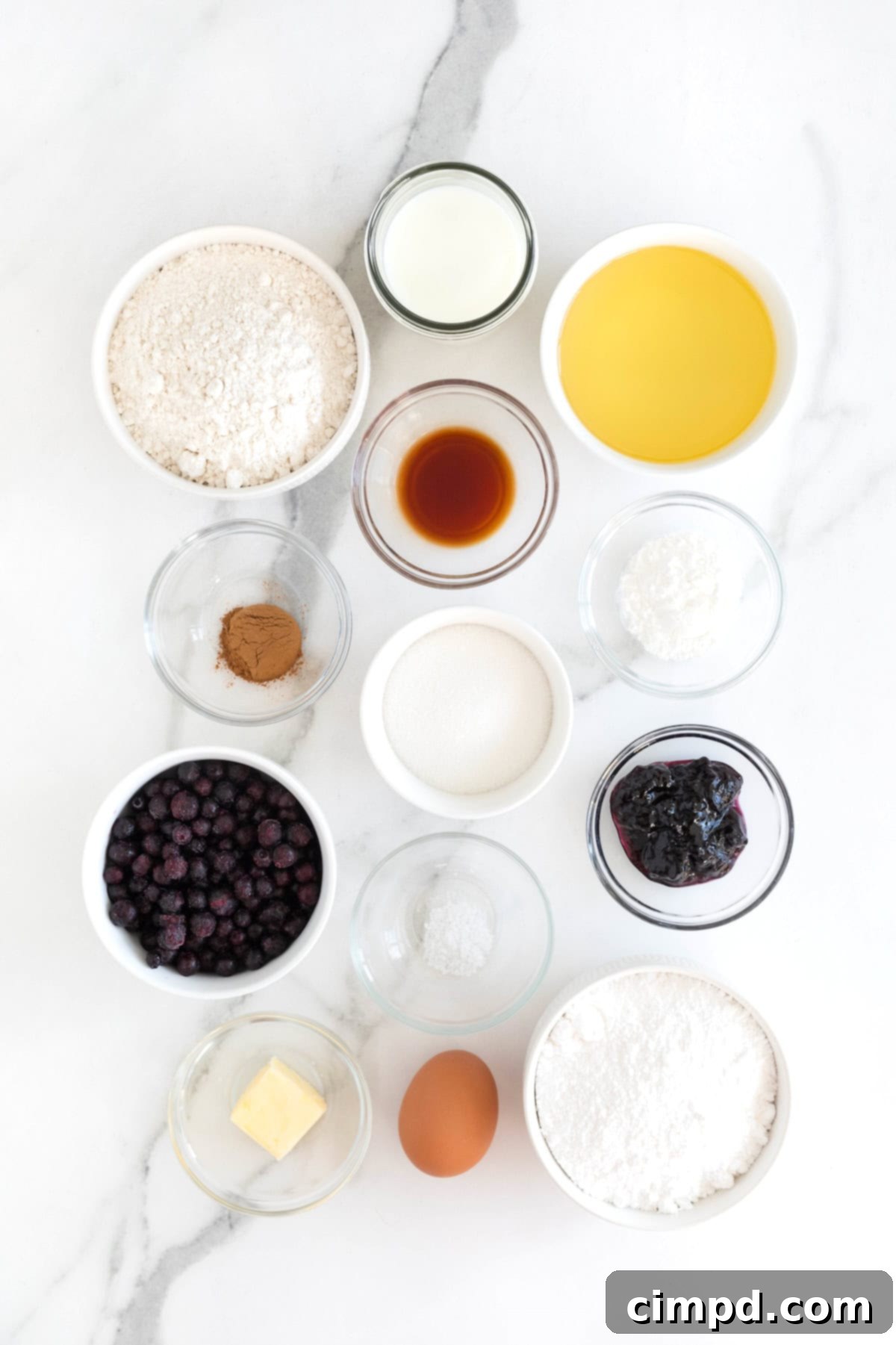 Individual ingredients for making homemade blueberry donuts, including flour, sugar, baking powder, cinnamon, salt, blueberries, milk, egg, vanilla, melted butter, and blueberry jam, arranged in small glass bowls on a white marble counter.