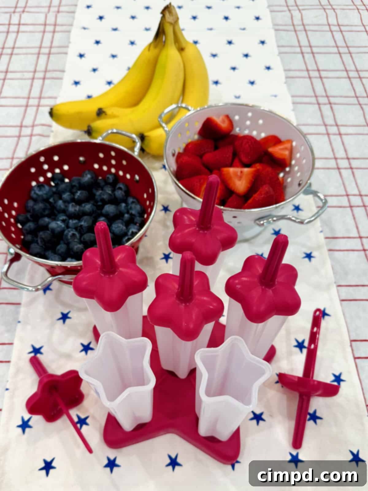 A neat arrangement of fresh strawberries, bananas, and blueberries, alongside a blender and popsicle molds, ready to make patriotic fruit pops.