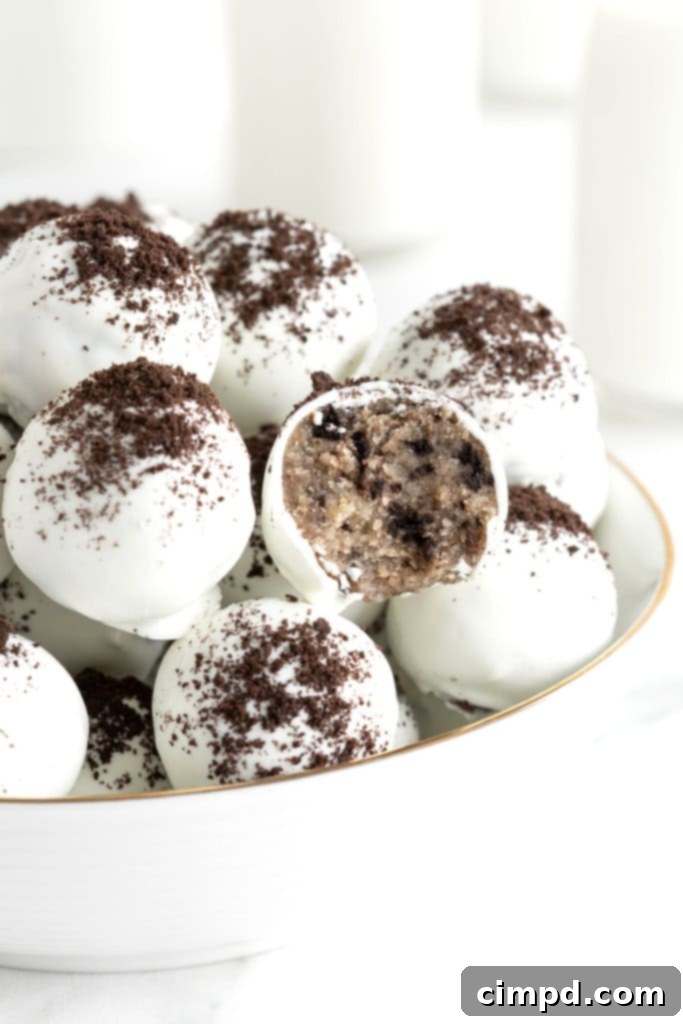 A large white serving bowl filled with cake balls coated in white coating and dusted with cookie crumbs. One of the cake balls has a bite out of it revealing the Oreo cookie cake inside.