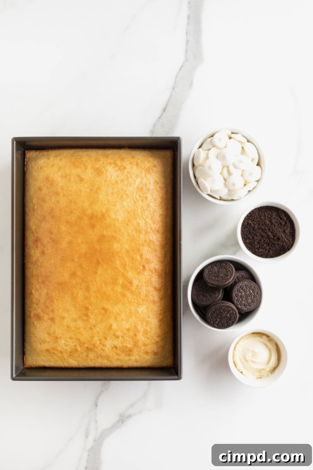 Ingredients to make Oreo cake balls - a 9x13 dark pan of white cake and Oreos, white candy coating, cookie crumbs in small white glass dishes on a white marble counter.