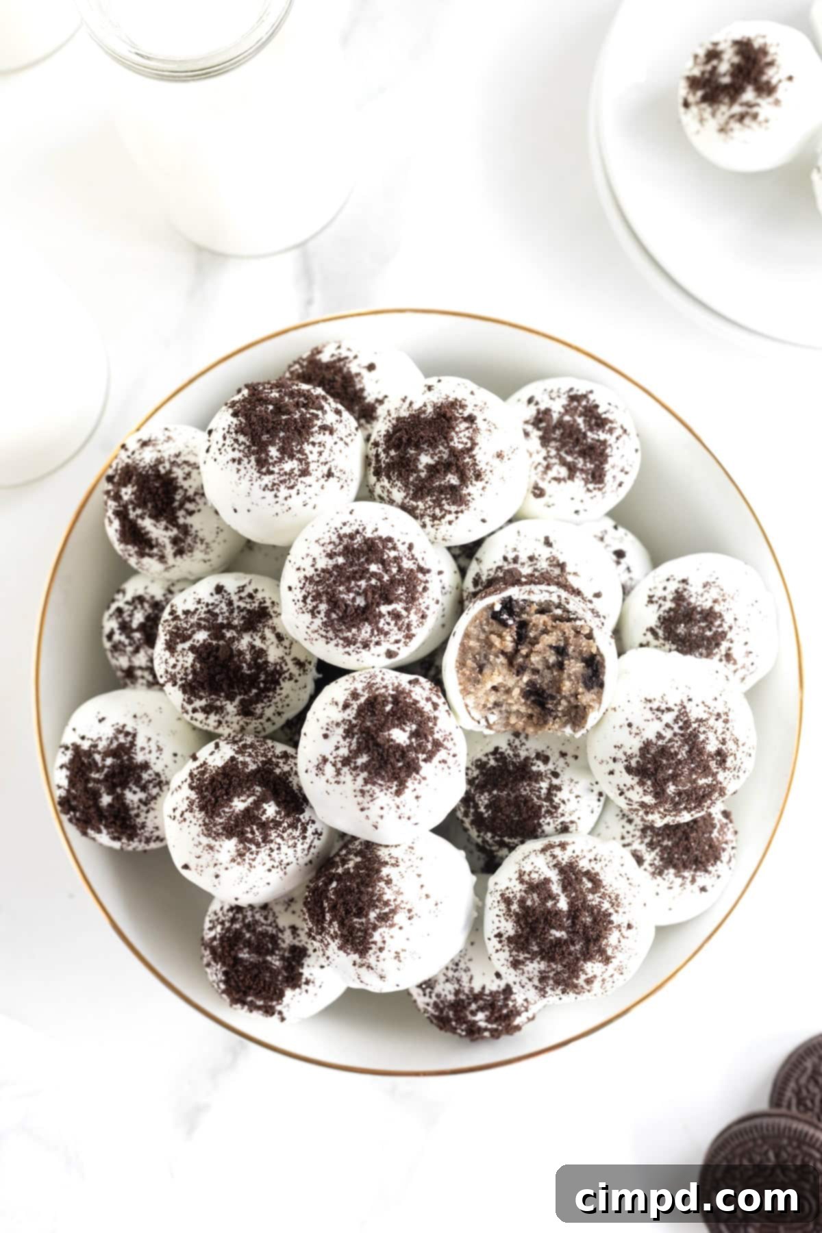 A large white serving bowl filled with cake balls coated in white coating and dusted with cookie crumbs. One of the cake balls has a bite out of it revealing the Oreo cookie cake inside.