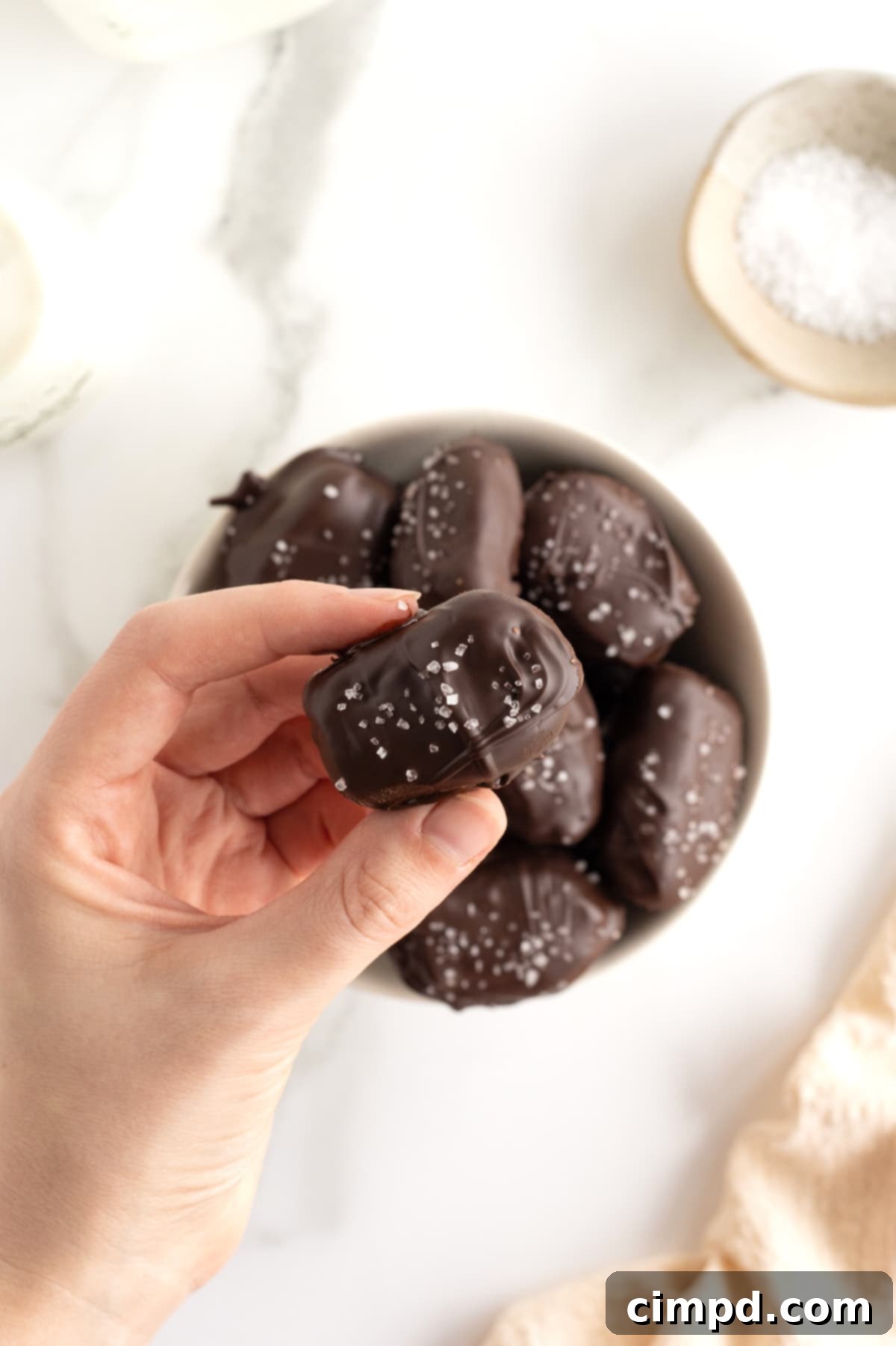 Salted Dark Chocolate Peanut Butter Stuffed Dates 5 A bowl of chocolate covered dates sprinkled with sea salt and a hand holding one of the dates between two fingers.