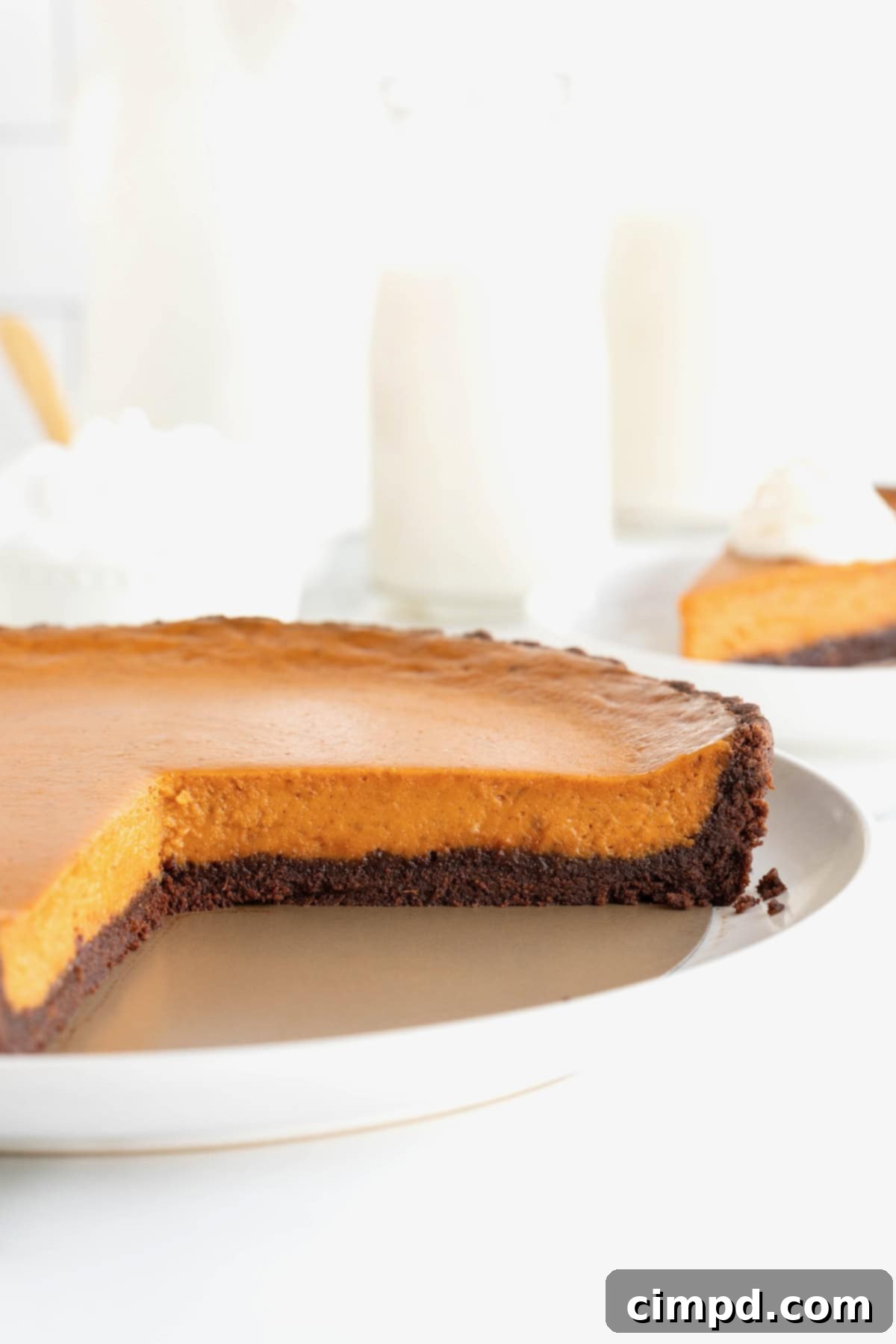 A round Dark Chocolate Pumpkin Tart, showcasing its robust chocolate crust, sits on a white serving plate. A perfectly cut wedge is missing, indicating it's already being enjoyed, leaving a tempting view of the creamy pumpkin interior.