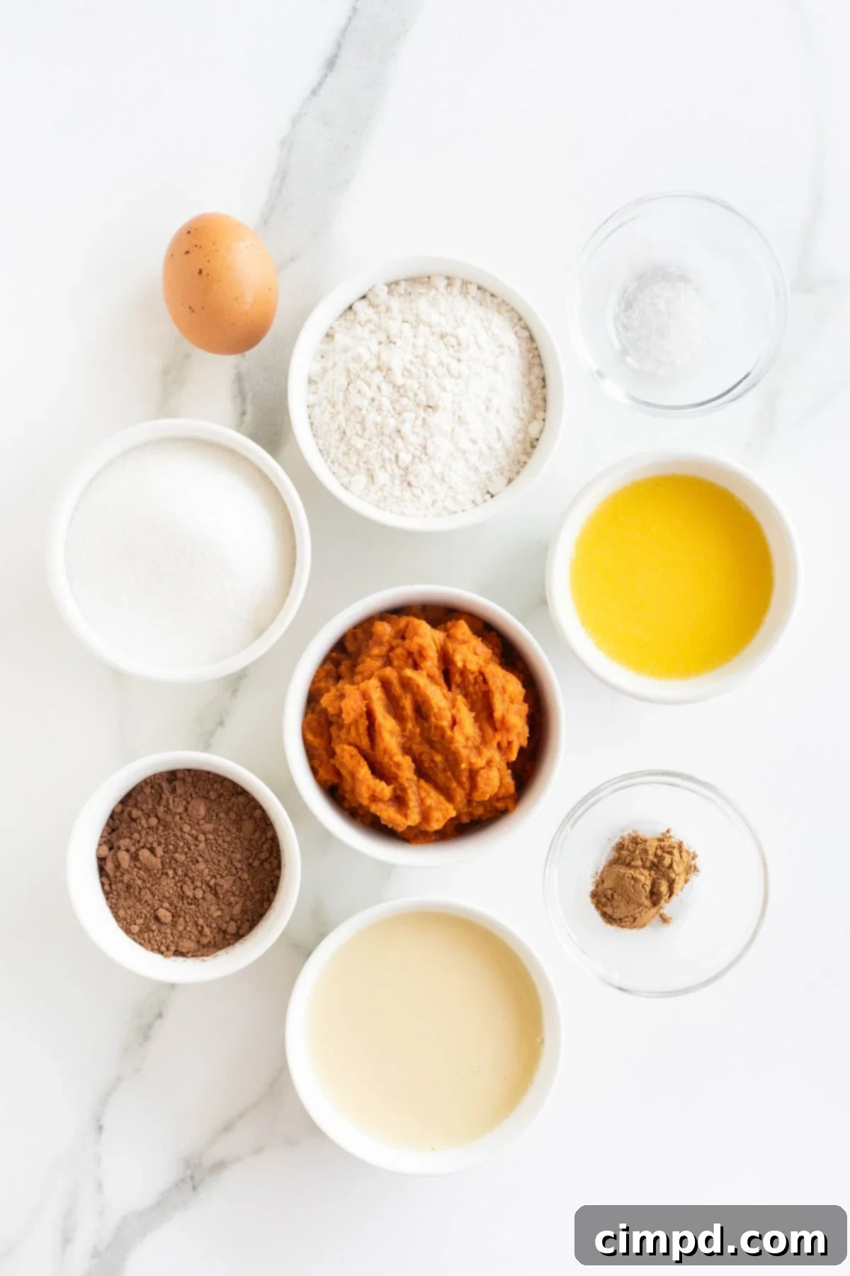 A curated display of ingredients for making a dark chocolate pumpkin tart, neatly arranged in small white glass dishes on a pristine white marble counter. The components are clearly visible, ready for baking.