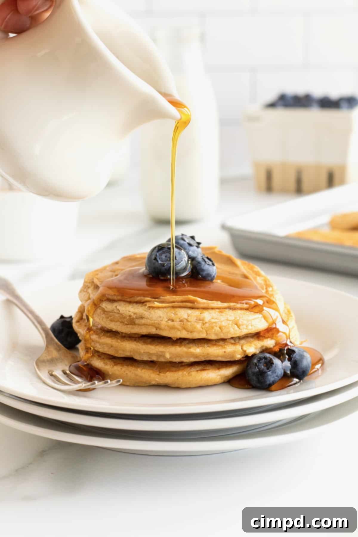 Simple 5-Ingredient Flourless Protein Pancakes 2 A beautifully stacked trio of golden-brown flourless protein pancakes, generously drizzled with maple syrup and adorned with a handful of fresh, vibrant blueberries. A spoonful of creamy peanut butter melts enticingly between the layers.