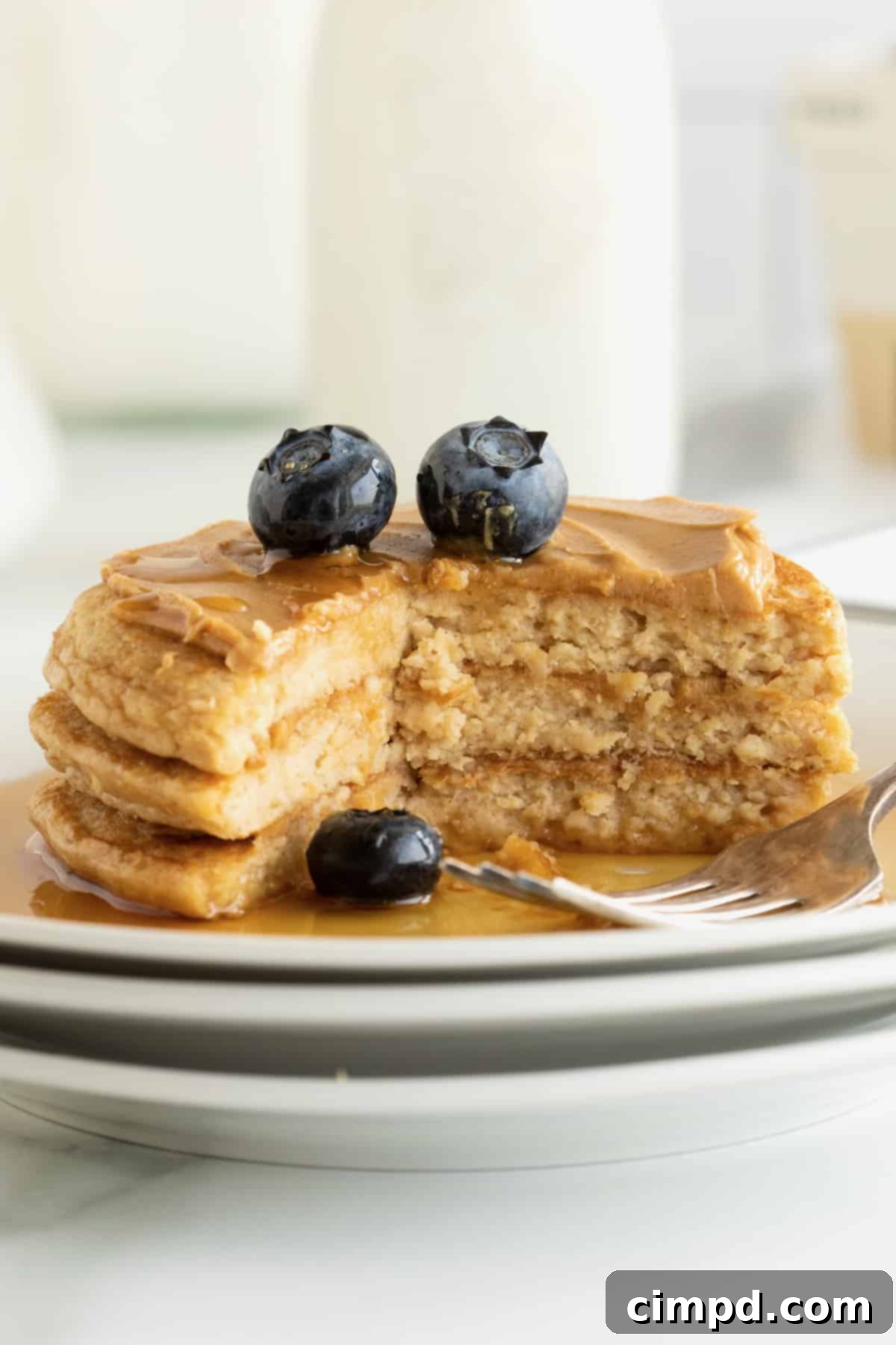 Simple 5-Ingredient Flourless Protein Pancakes 3 A close-up shot of a stack of three protein pancakes, artfully smeared with peanut butter and garnished with fresh blueberries and a generous pour of golden maple syrup, ready to be enjoyed.