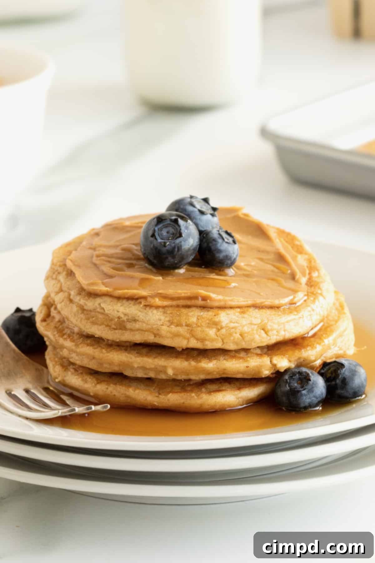 Simple 5-Ingredient Flourless Protein Pancakes 5 A tempting stack of three flourless protein pancakes, smothered in creamy peanut butter and adorned with fresh blueberries and a glossy drizzle of maple syrup, presented on a rustic wooden surface.
