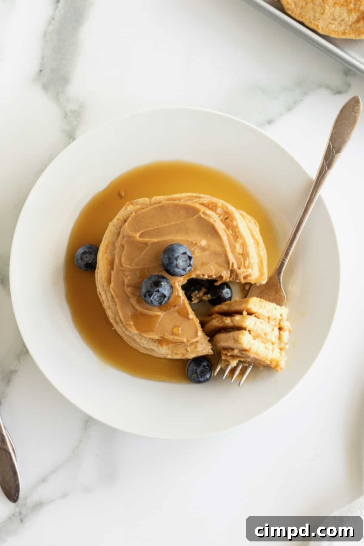 Simple 5-Ingredient Flourless Protein Pancakes 6 A vibrant and appetizing close-up of a stack of three protein pancakes, generously slathered with peanut butter and topped with an abundance of fresh blueberries and a golden stream of maple syrup.